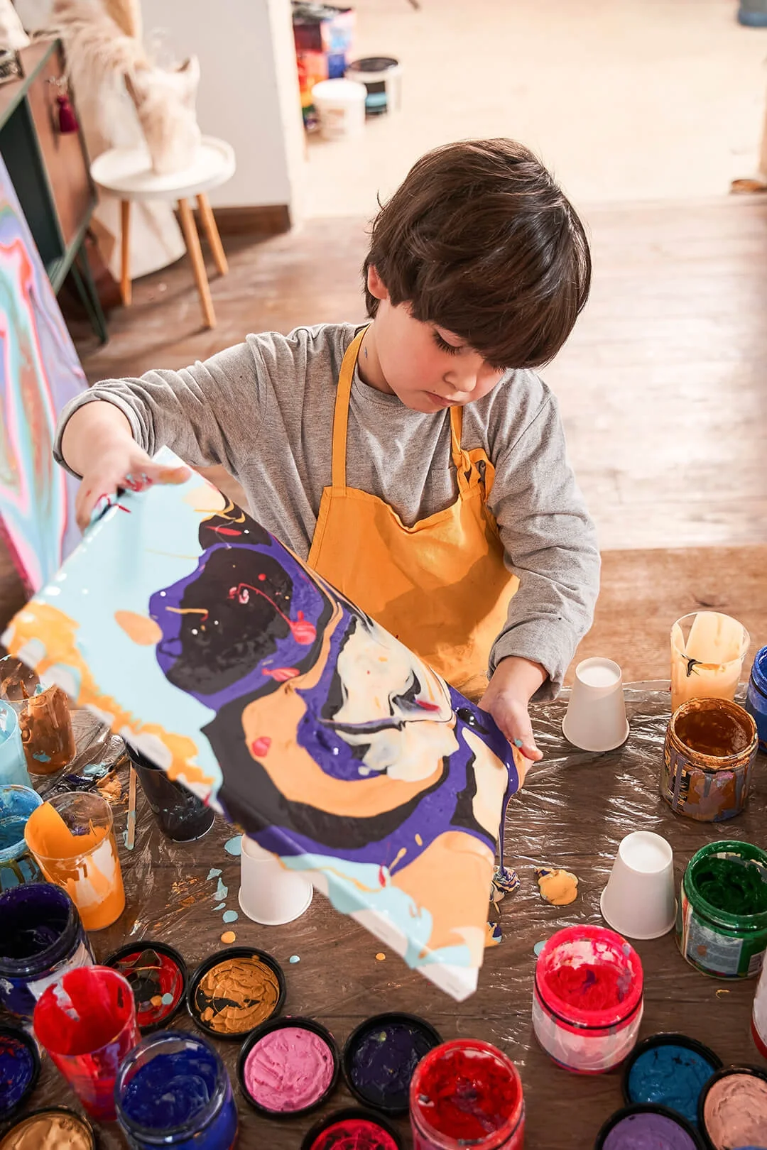 Young boy tilting canvas to create flowing, colorful paint design on canvas.