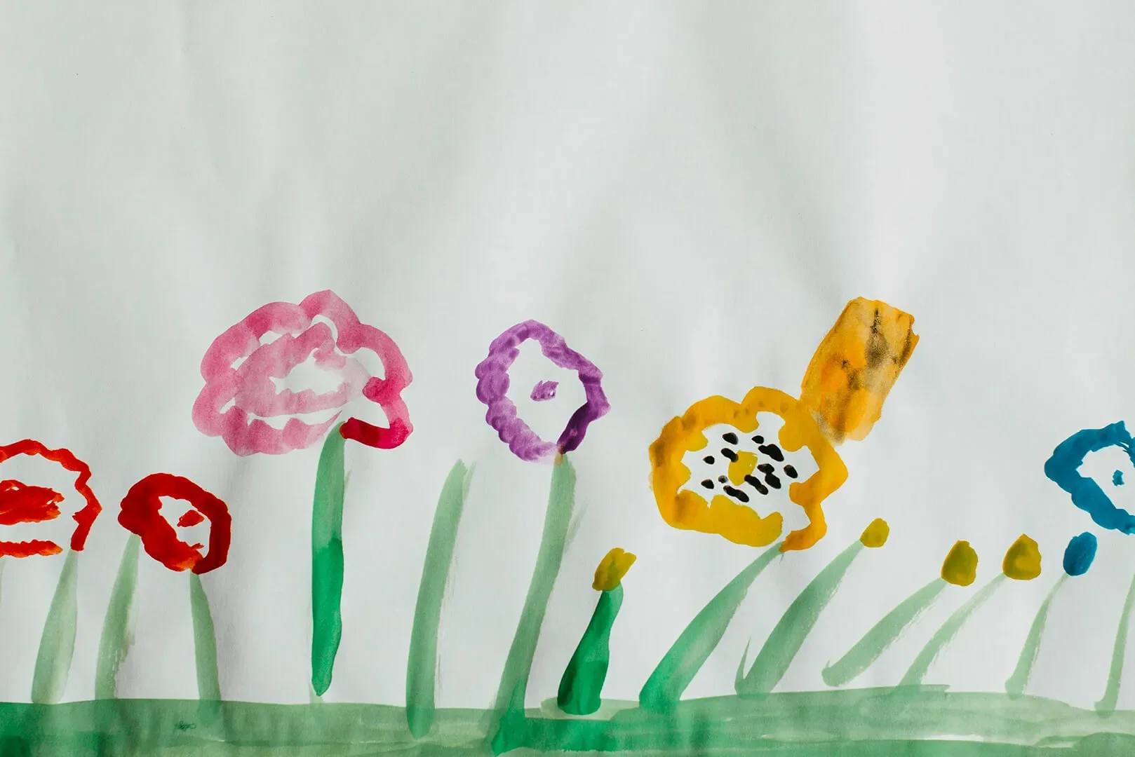 Watercolor painting of simple flowers in red, pink, purple, yellow, and blue with tall green stems on folded white paper.