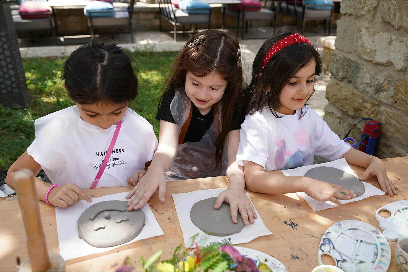 Three young girls making clay art faces in an outdoor setting.