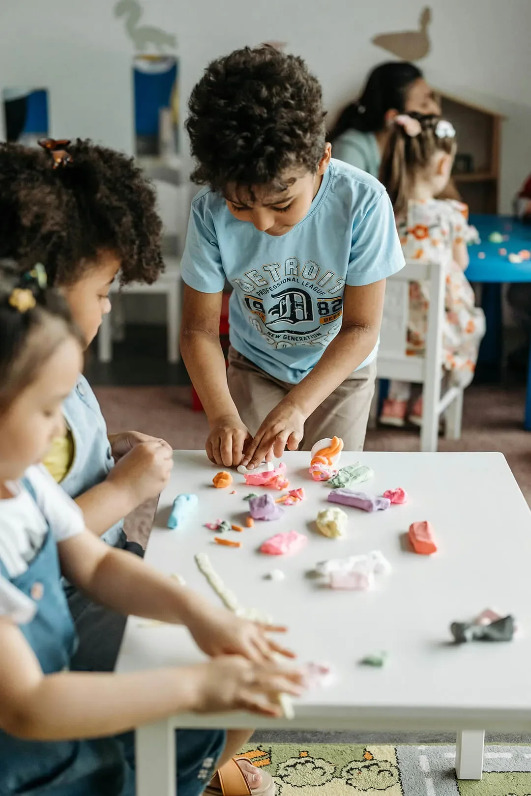 Three young children working together and crafting with colored clay at white work table.