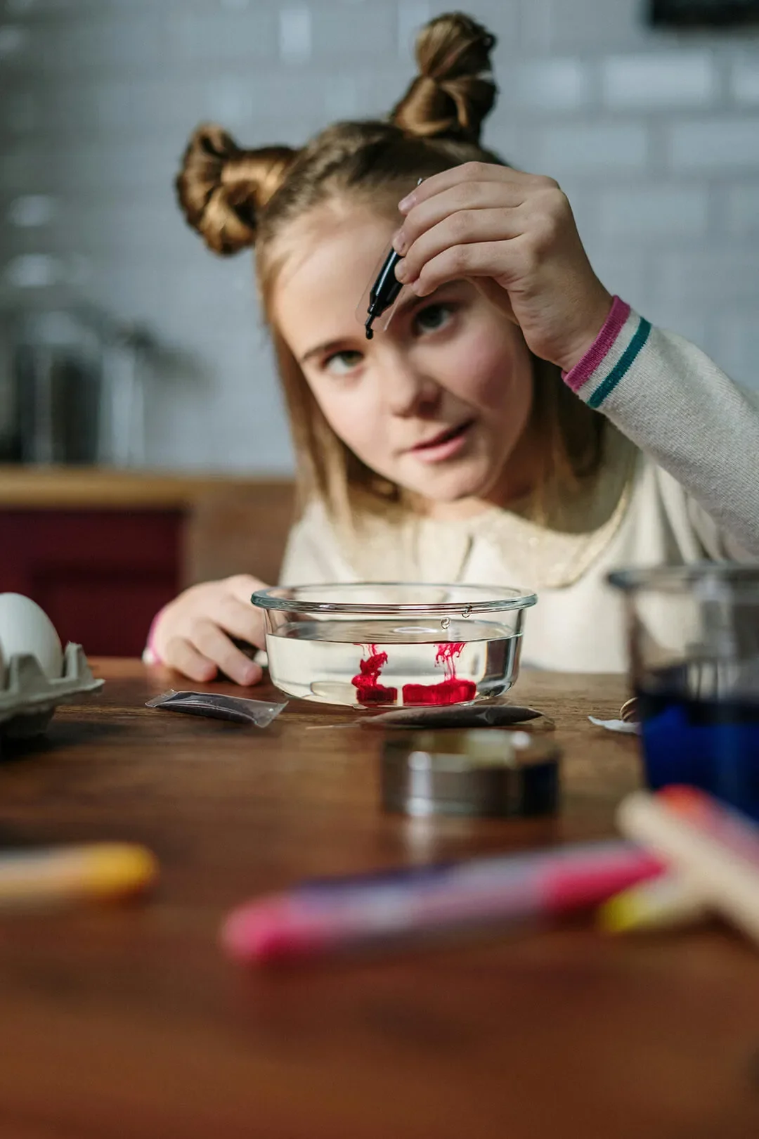 Young girl drops colored dye into bowl full of clear liquid and observes from wooden table top.