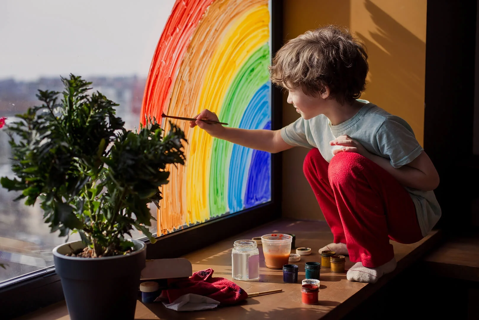 Young boy painting rainbow color arch onto glass window.