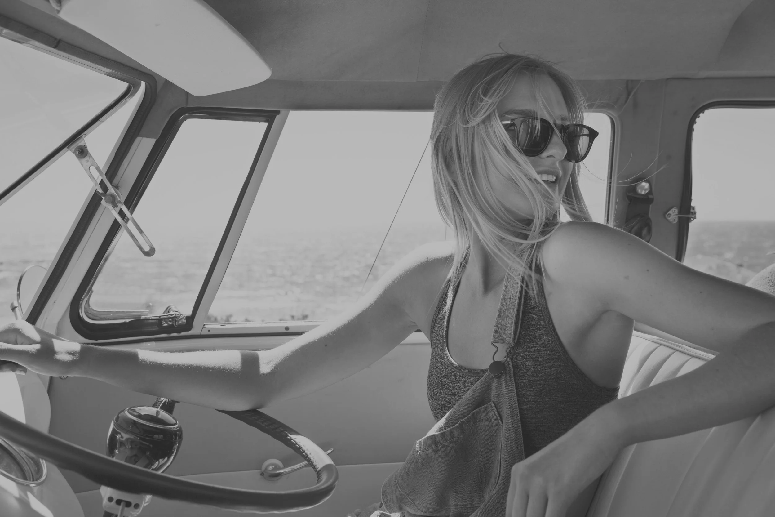 A woman with blonde hair and sunglasses smiling while sitting in the driver's seat of a vintage vehicle, with the ocean visible through the open window.