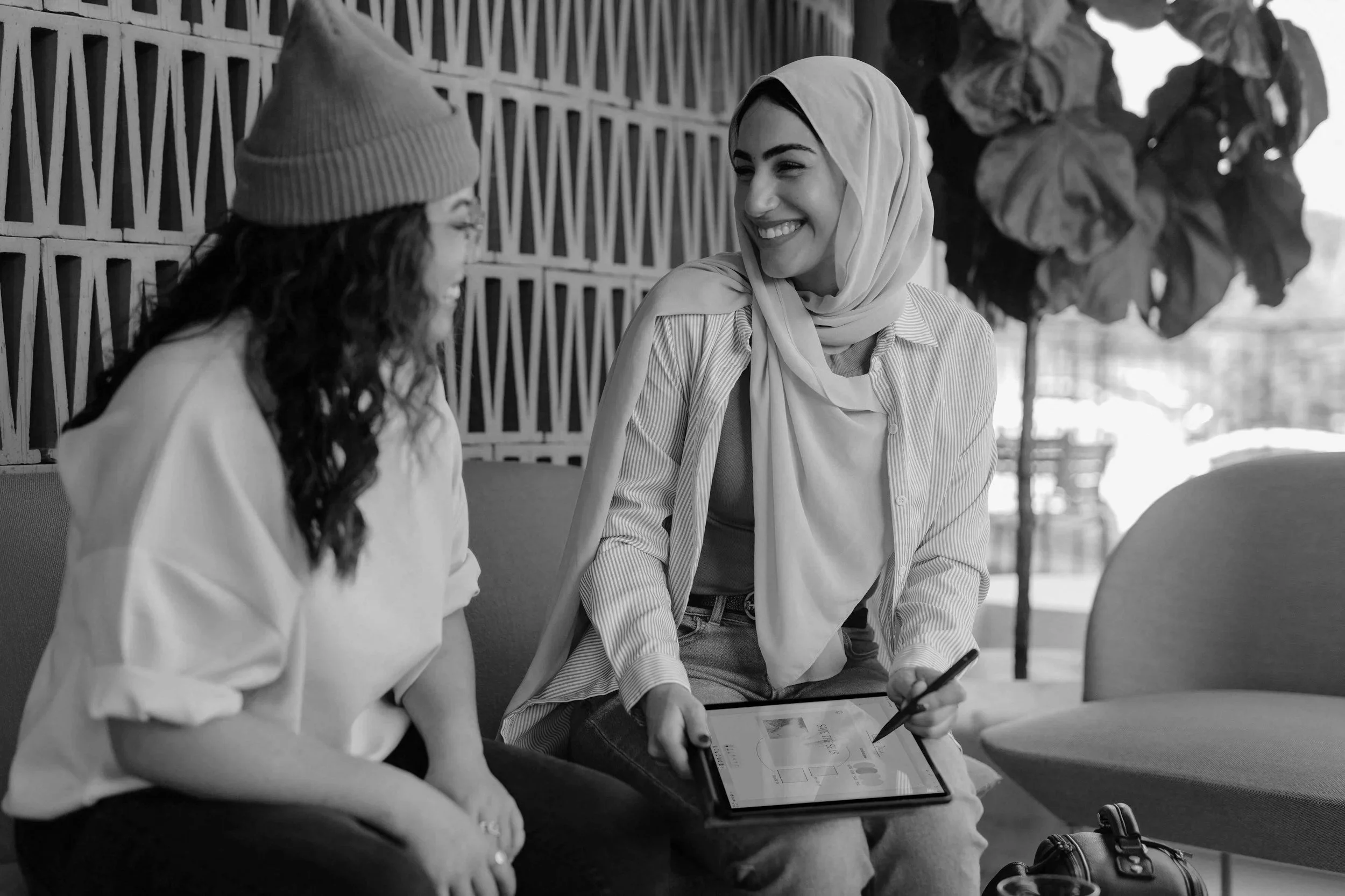 Two women are sitting on a couch, engaging in conversation. One is wearing a beanie and a loose shirt, the other is wearing a headscarf and a striped shirt, and she is holding a tablet with a stylus, smiling.