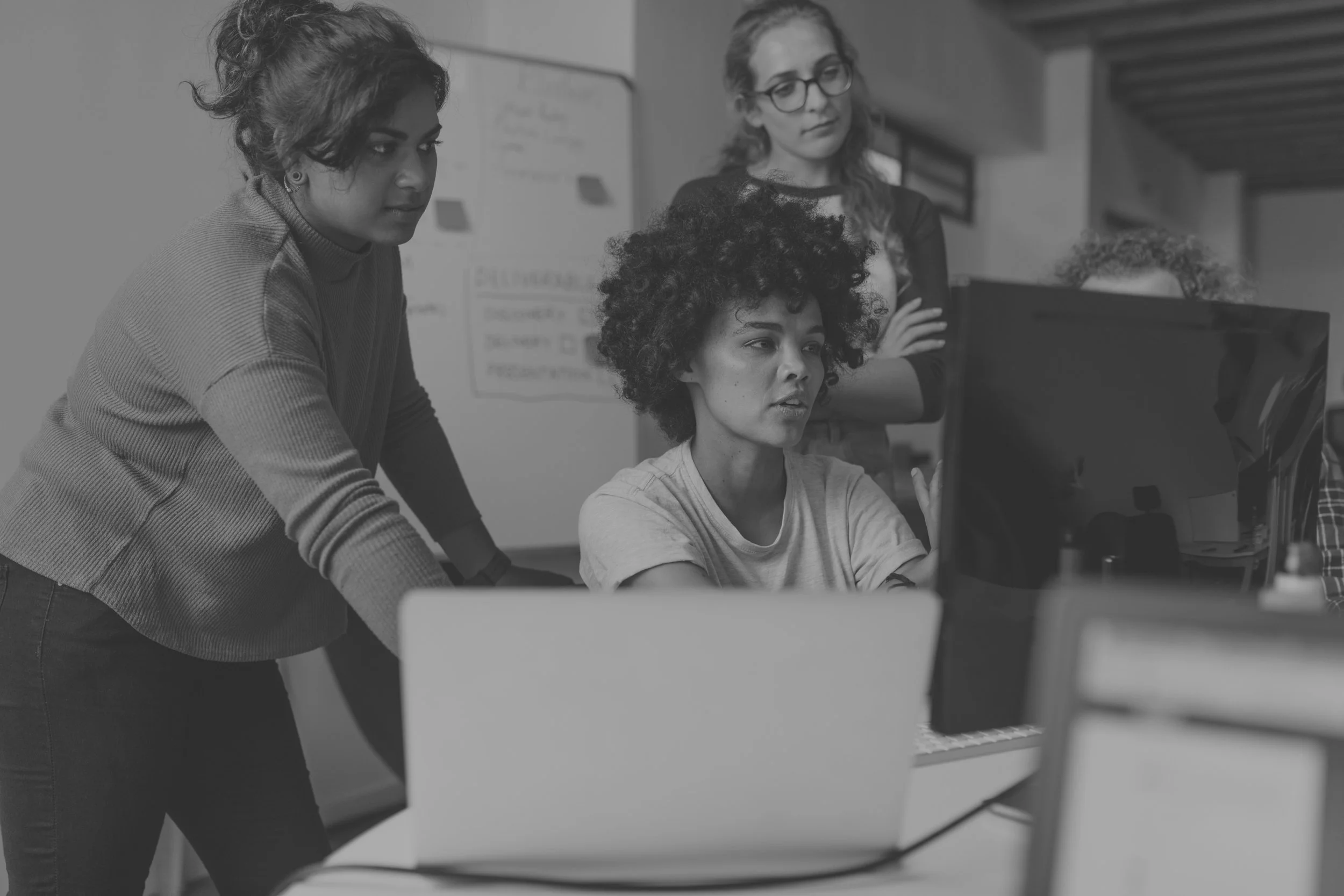 Four women gathered around a computer in an office setting, focused on the screen, engaged in a discussion or collaboration.