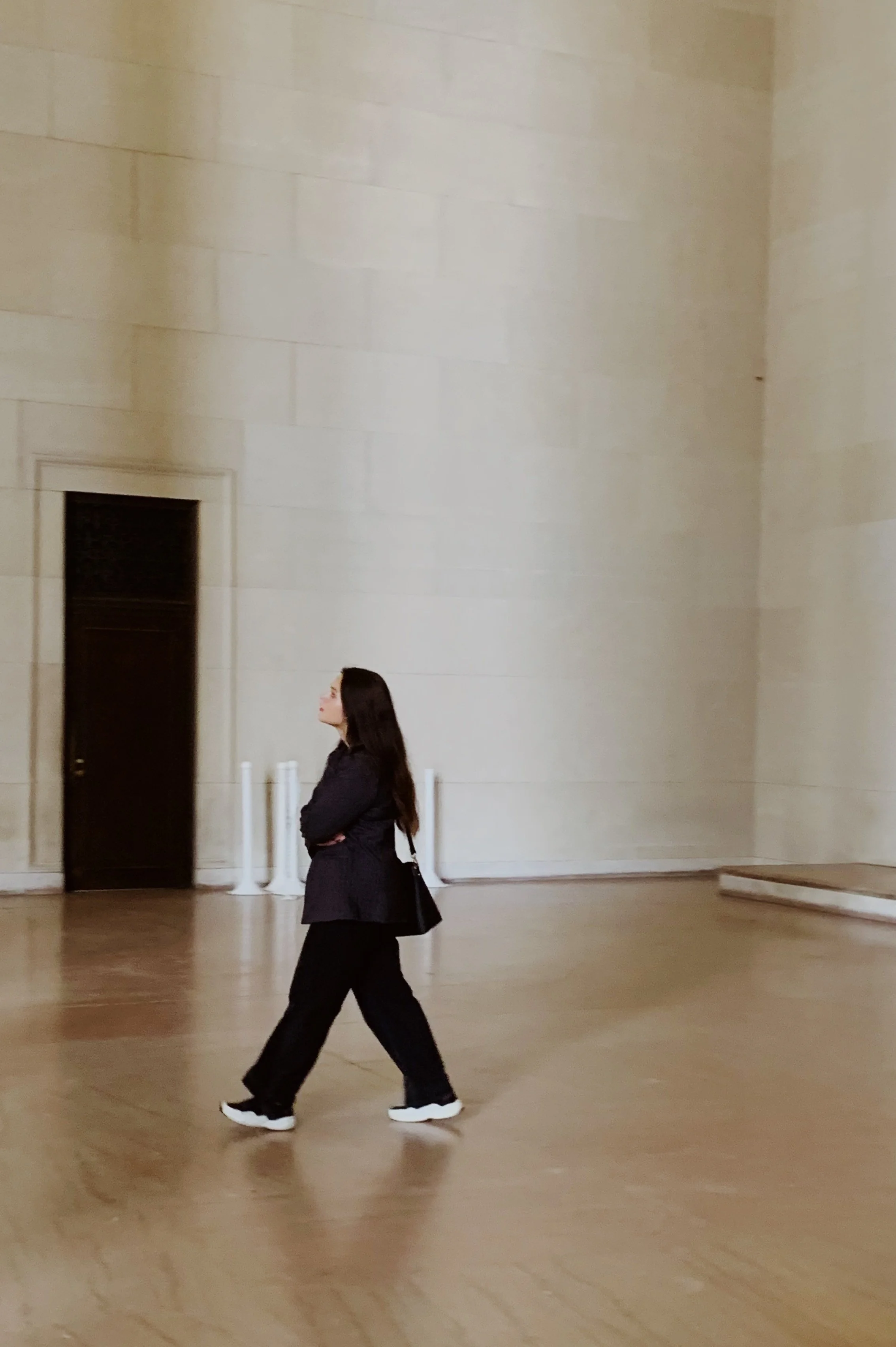 A woman dressed in black walking across a large, empty space with beige walls and wooden flooring.
