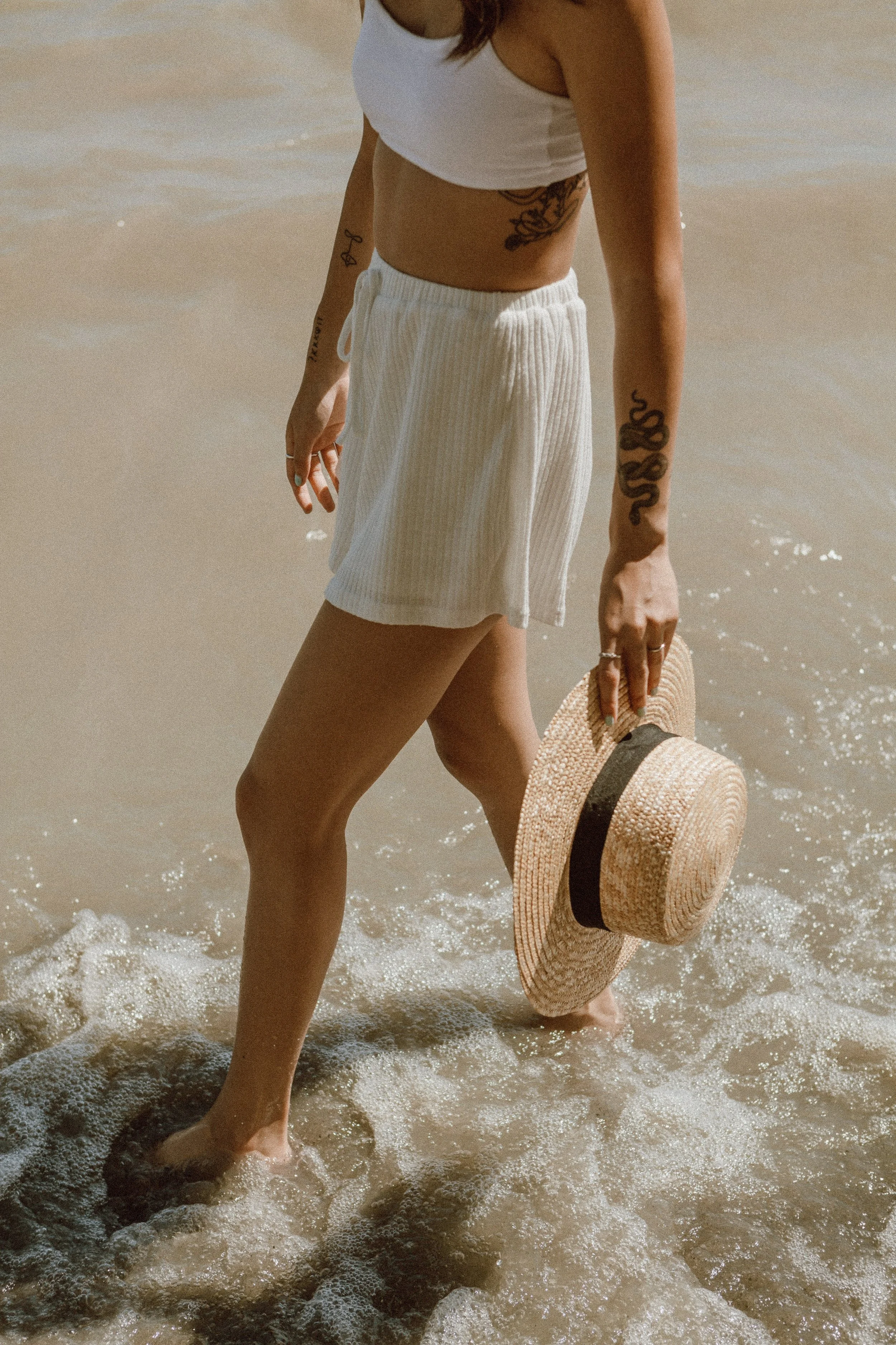 A woman standing in shallow water at the beach, holding a straw hat, wearing a white crop top and shorts, with tattoos on her arms and torso.
