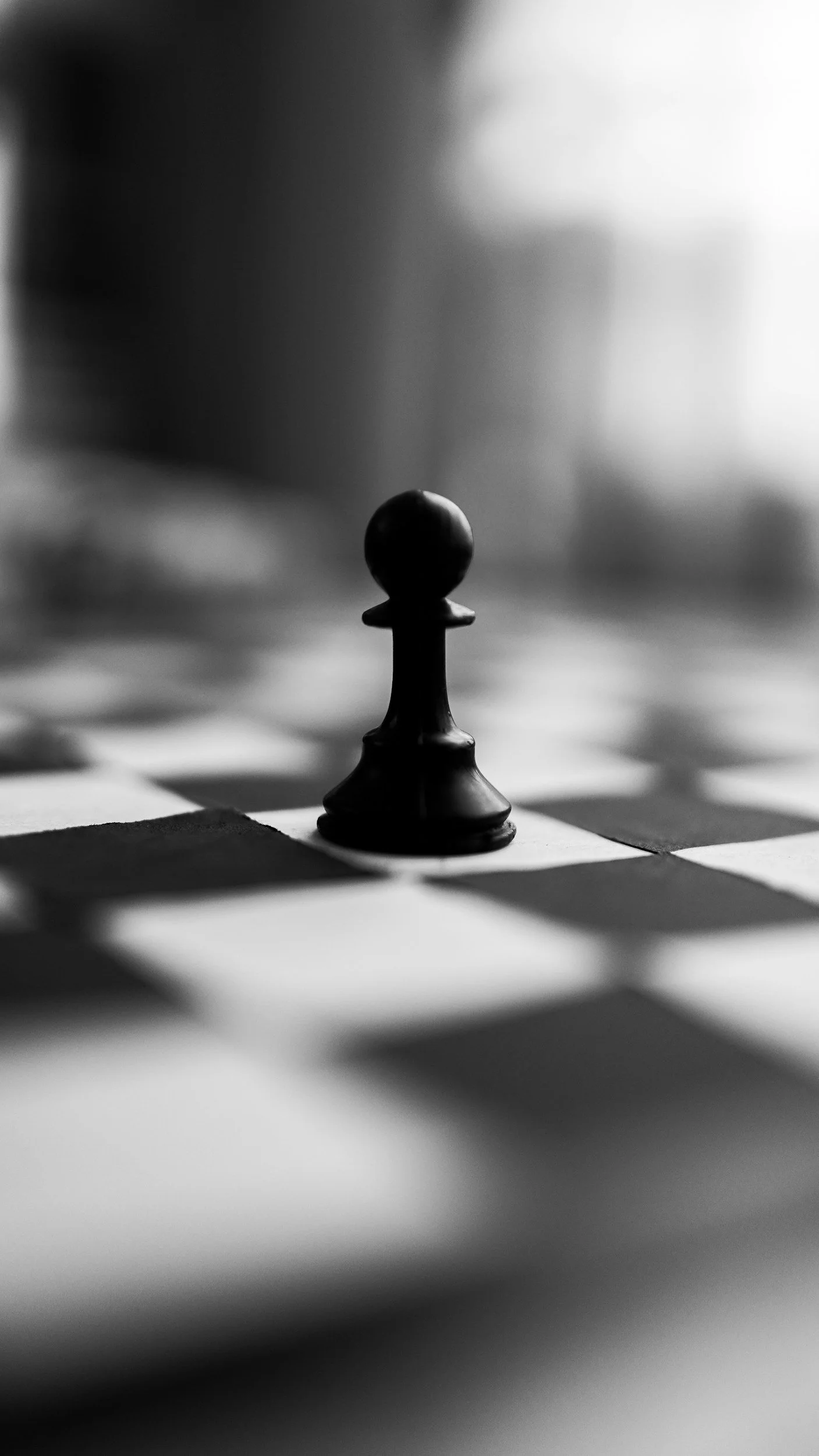 Close-up of a single black pawn on a chessboard, in black and white. The focus is on the pawn, with the background and other squares blurred.