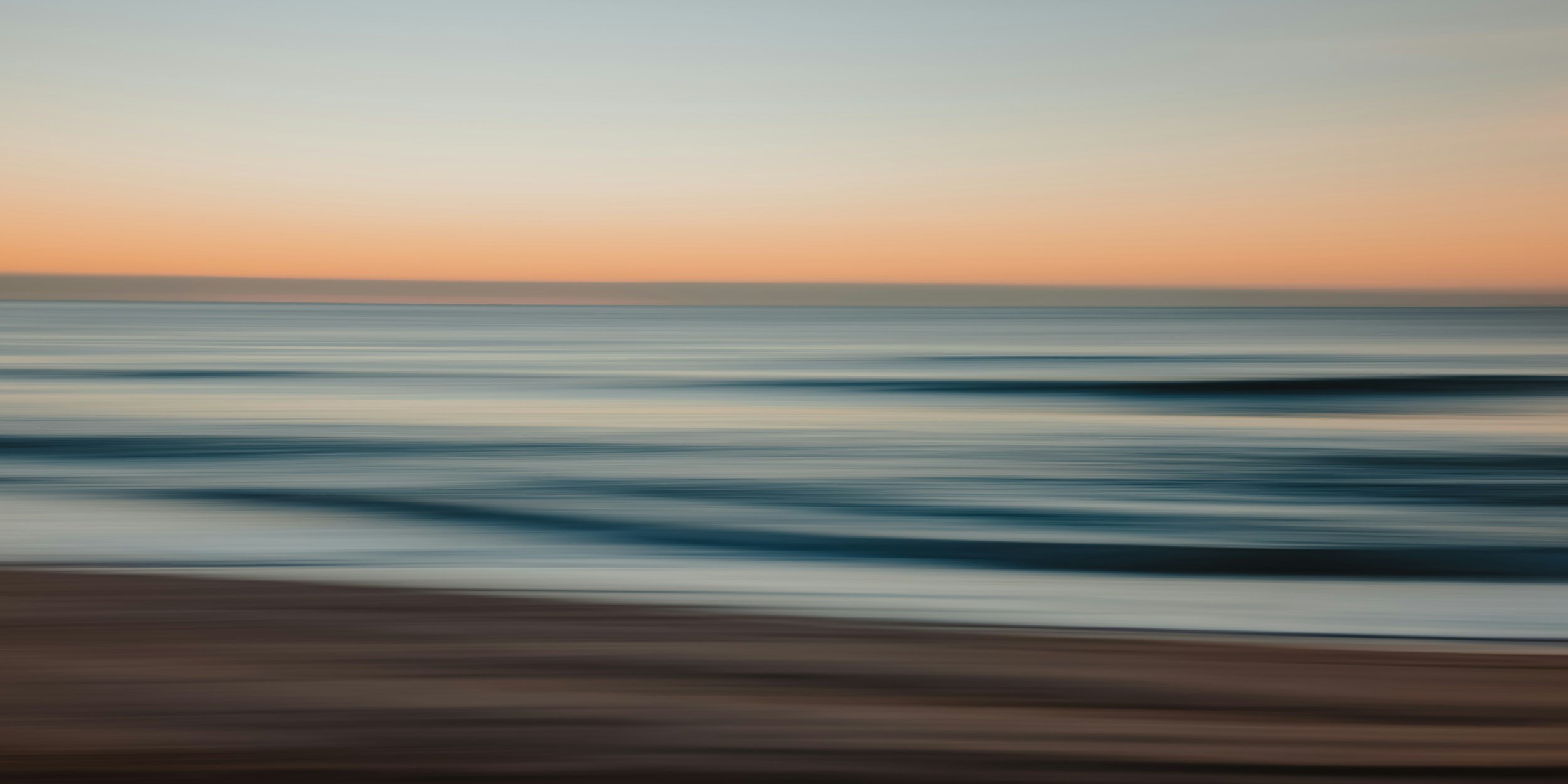 Blurred image of a beach at sunset with waves crashing on the shore and a colorful sky.