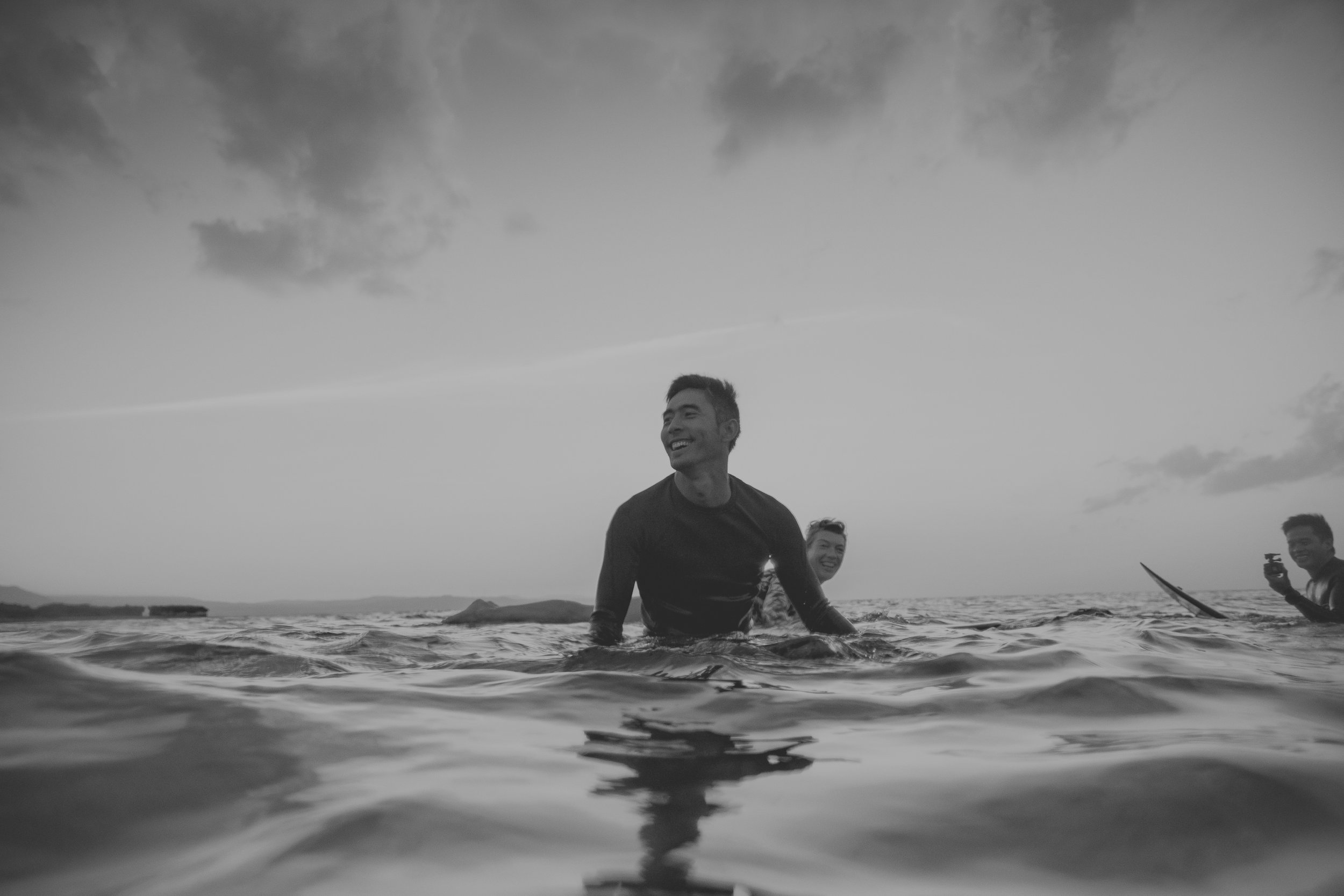 People enjoying their time in the ocean, smiling and taking photos, with a cloudy sky in the background, black and white photo.