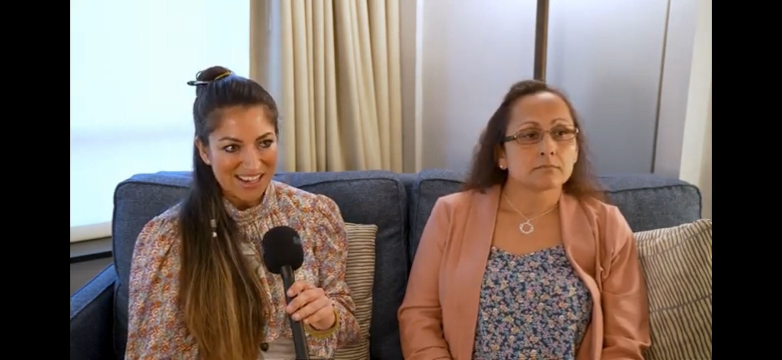 Two women sitting on a gray couch in a room with beige curtains and wall paneling, one holding a microphone and smiling, the other looking serious and wearing glasses.