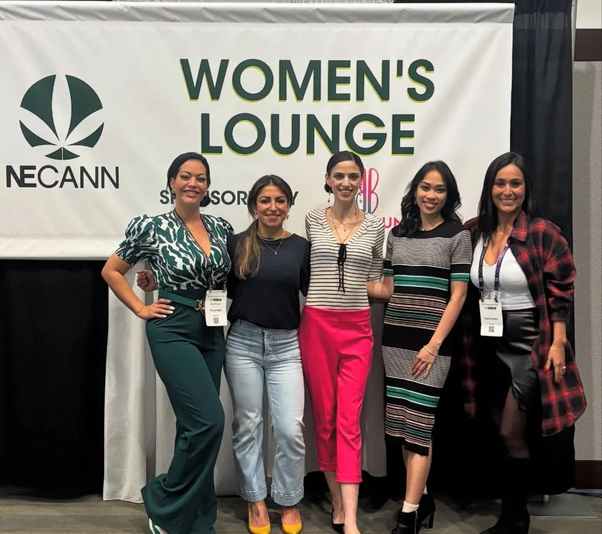 Five women standing together at a Women’s Lounge event, smiling, in front of a large NECANN banner. They are dressed in various fashionable outfits, some with name tags.