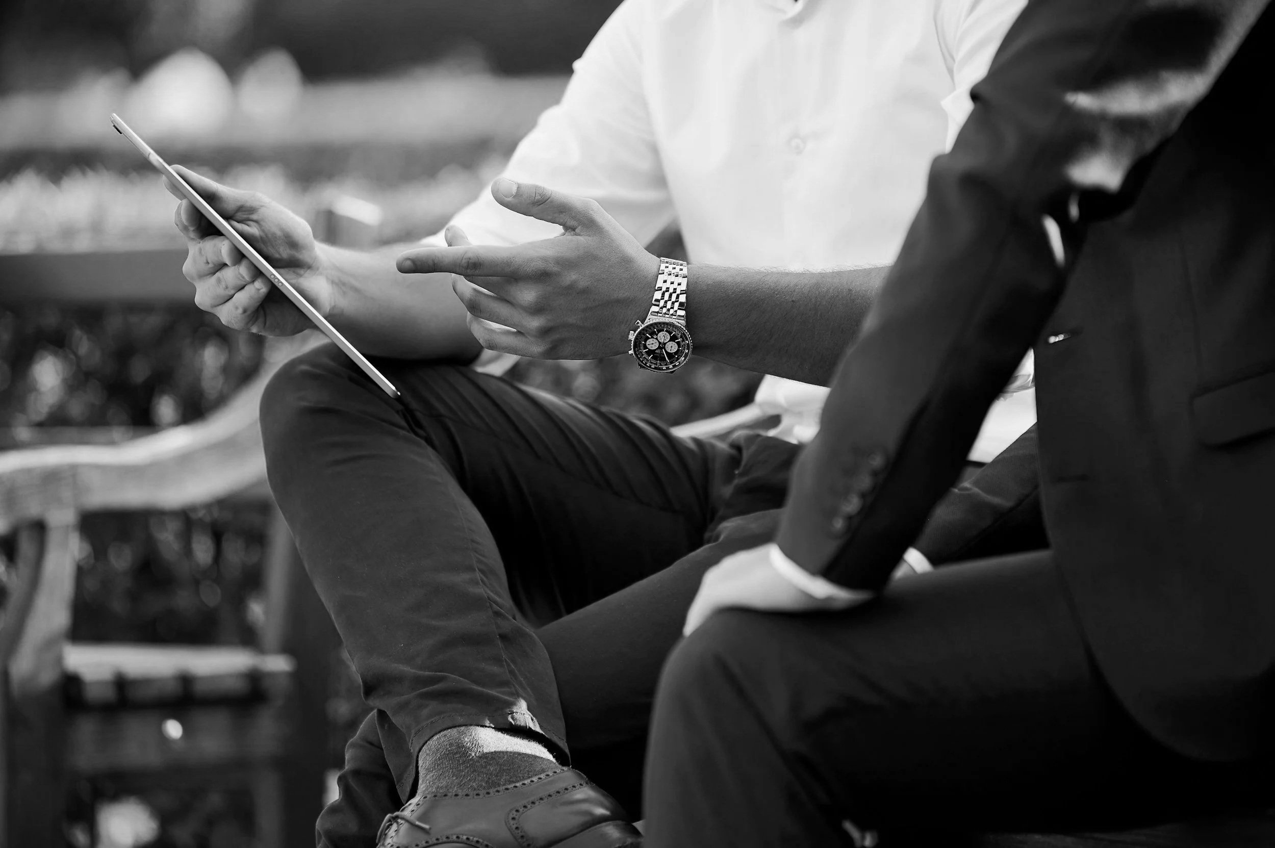 Two men sitting and discussing, one holding a tablet and pointing at it, both dressed in formal business attire, one wearing a watch, outdoors.