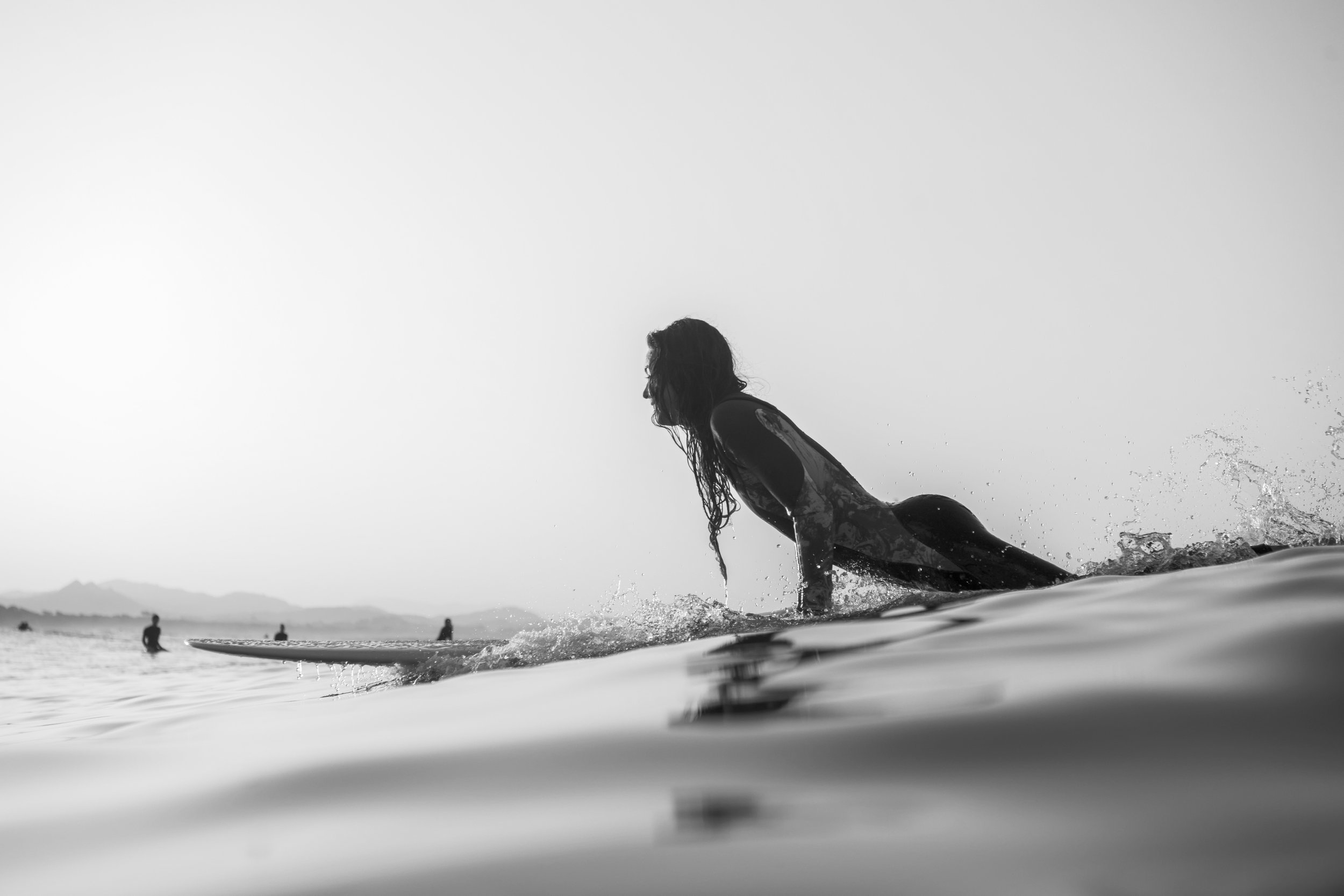 A black and white photo of a woman on a surfboard in the water, with other surfers in the distance and a mountain range on the horizon.