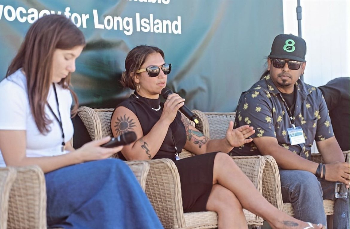 Three individuals sitting on wicker furniture at an outdoor event, with a blue banner behind them that reads 'Occupy for Long Island'. The woman in the middle is speaking into a microphone, wearing sunglasses, a black dress, and has tattoos on her ar
