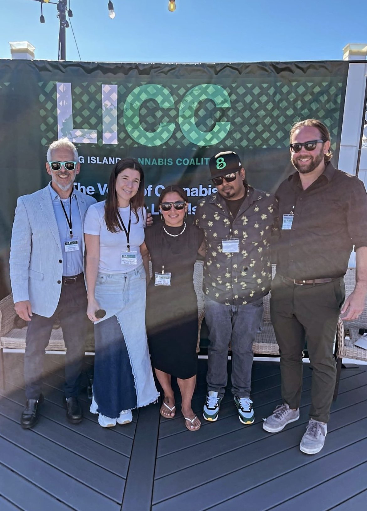 A group of five people standing outdoors in front of a banner that reads "High Cannabis Coalition" with two of them holding microphones. The group is smiling and wearing sunglasses, posing for a photo on a wooden deck with string lights overhead.