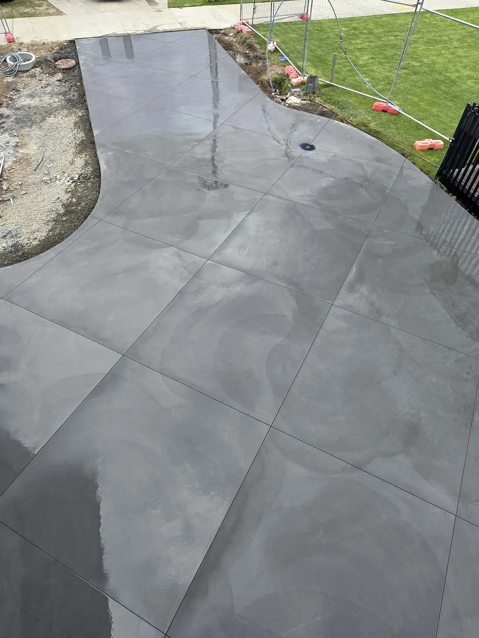 Recently poured and cleaned concrete patio with a polished gray surface, situated near a grassy area and construction fencing.