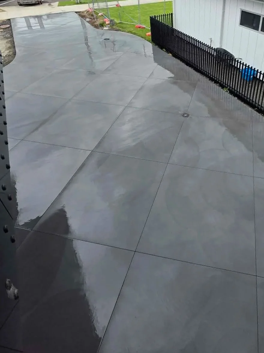A wet, shiny concrete patio area with reflections from the cloudy sky above, bordered by a black fence and a small grassy yard with orange safety barriers in the background.