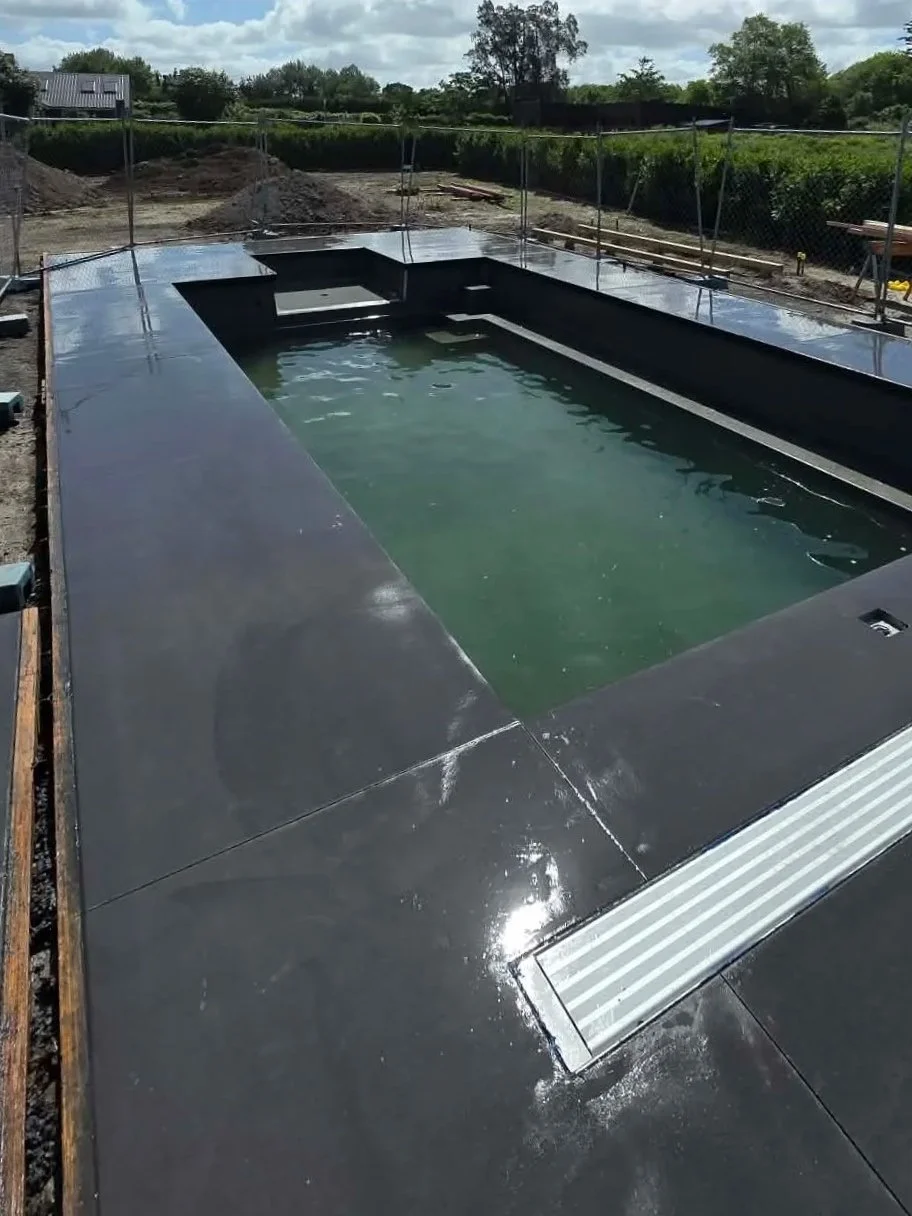 Empty rectangular swimming pool under construction with black lining, surrounded by a construction site fence and outdoor green trees and grass
