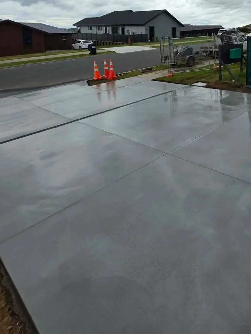 Newly poured wet concrete driveway with orange road cones at the edge, neighboring houses and parked cars visible in the background on a cloudy day.