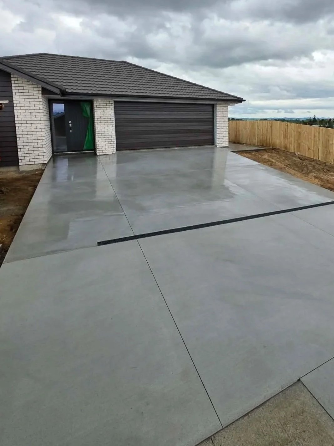 Newly poured concrete driveway in front of a house with a gray garage door, brick exterior, and a wooden fence on the side, under a cloudy sky.