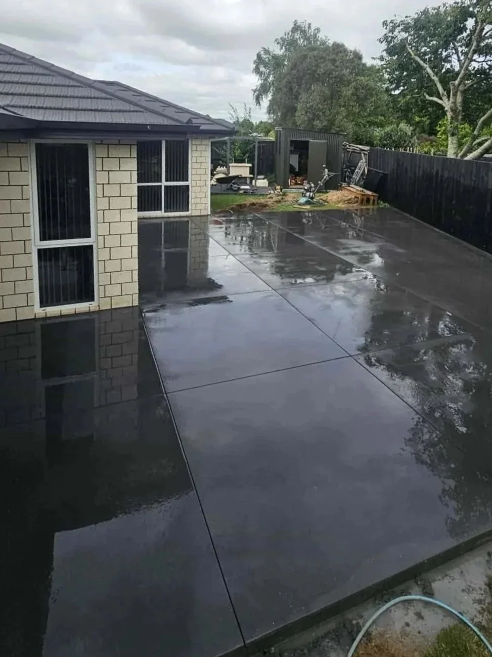 A freshly poured black concrete patio outside a house on a cloudy day, with some reflections visible on the wet surface, and a shed in the backyard.