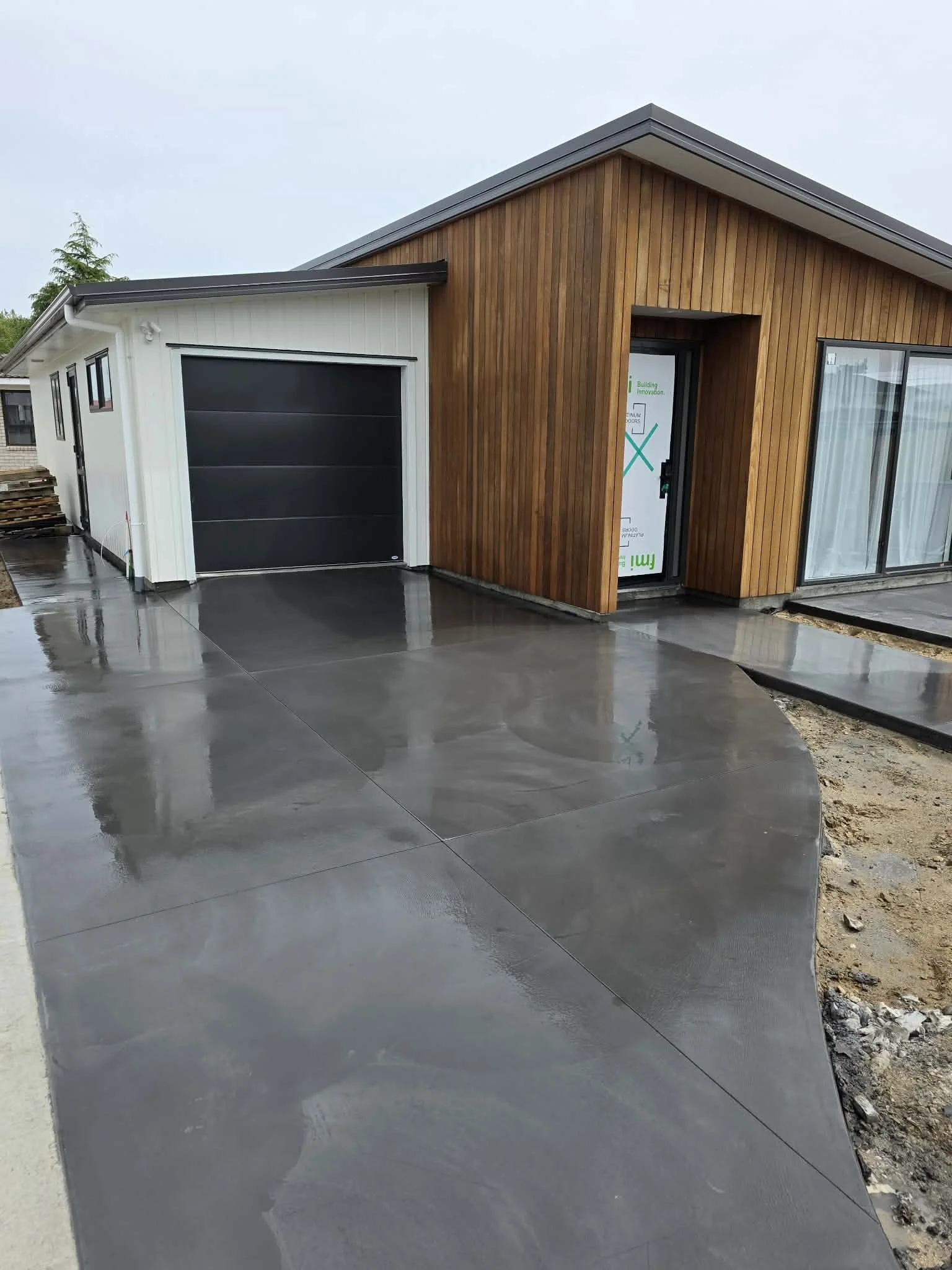 Newly constructed modern house with a black garage door, wood and white exterior, large sliding glass door, and a freshly poured wet concrete driveway.