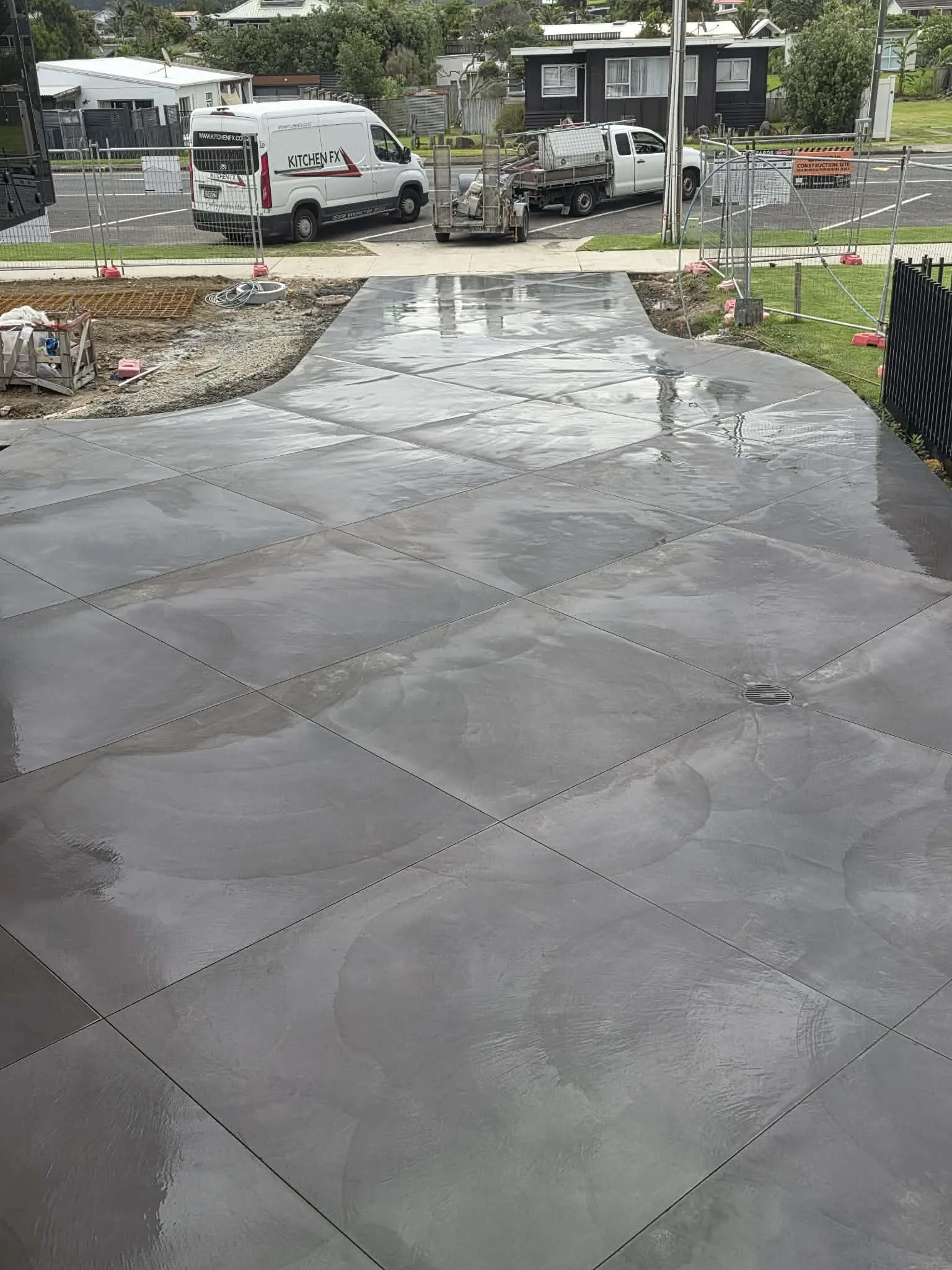Wet concrete patio with large square tiles in a residential area, construction site fencing, construction trucks, and houses in the background.