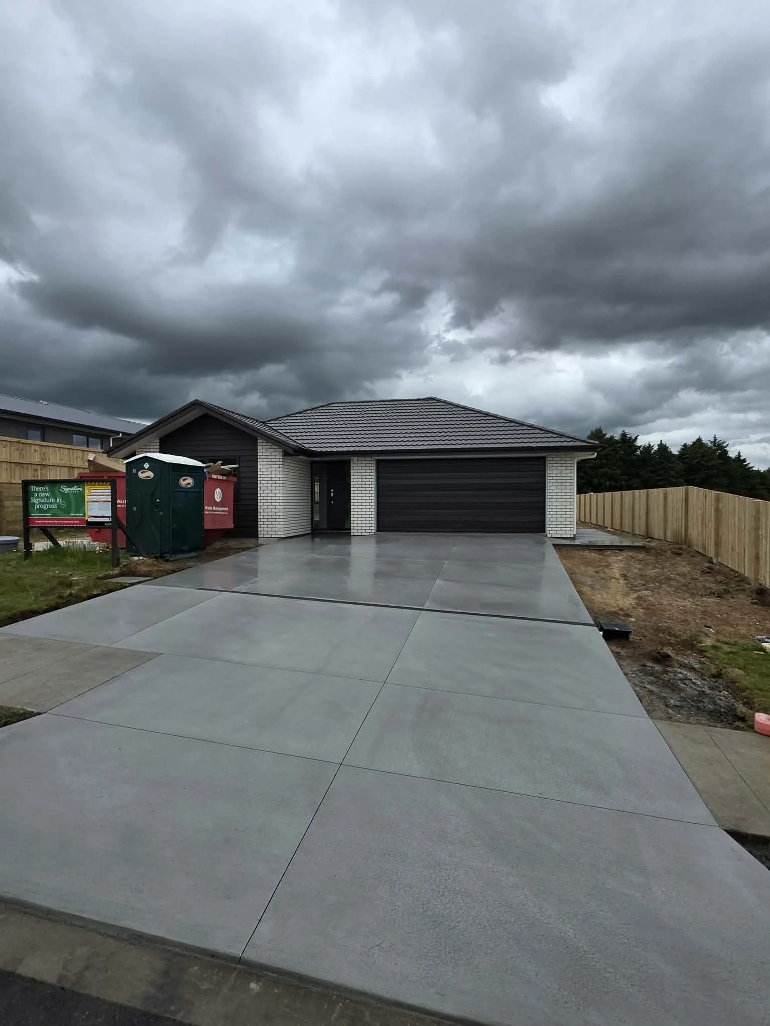Freshly paved concrete driveway in front of a house with a closed garage door, dark cloudy sky overhead, and new fence on sides.