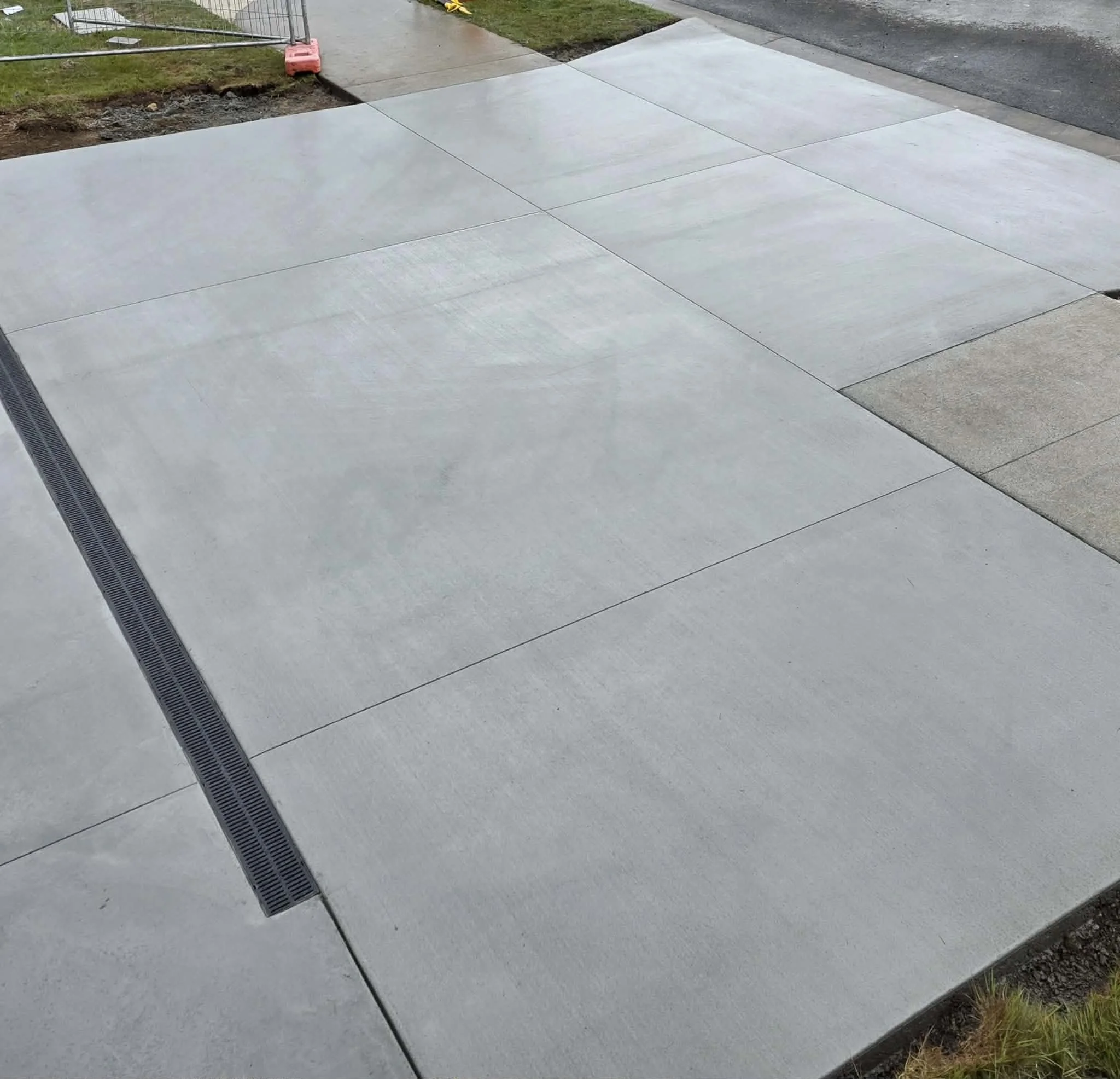 Concrete sidewalk with a drainage grate, partially wet and bordered by dirt and grass.