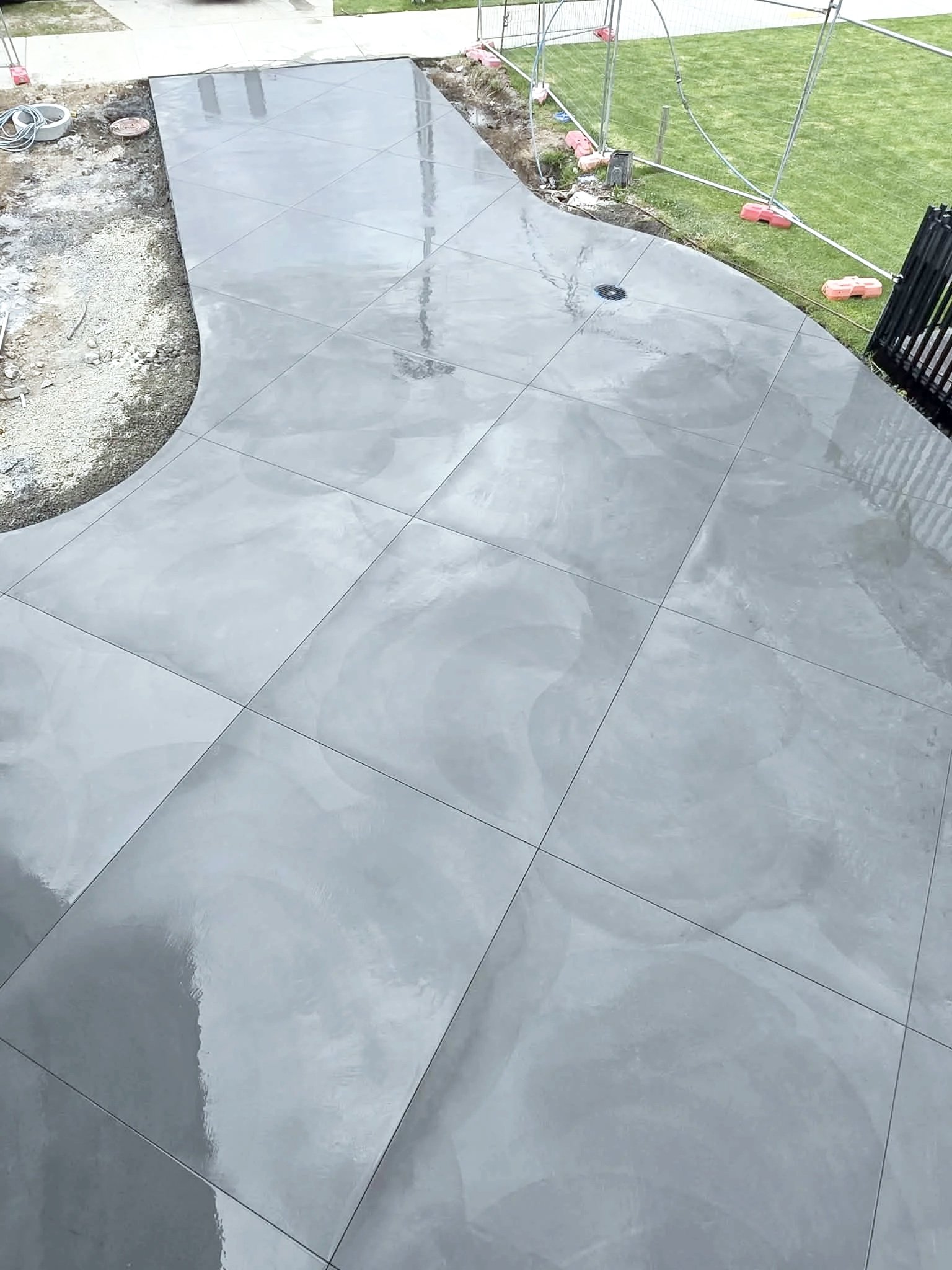 Freshly installed large gray tiles on a patio or walkway area next to a grass lawn and construction fencing.