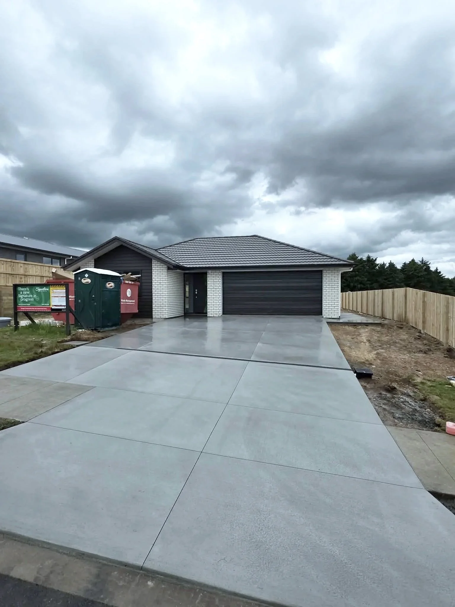 Newly constructed modern house with black garage door, white brick walls, and a freshly poured concrete driveway, under cloudy skies.