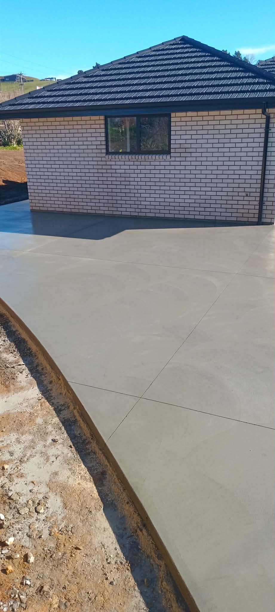 Newly poured concrete patio adjacent to a brick house with a black tiled roof and a large window, under a clear blue sky.