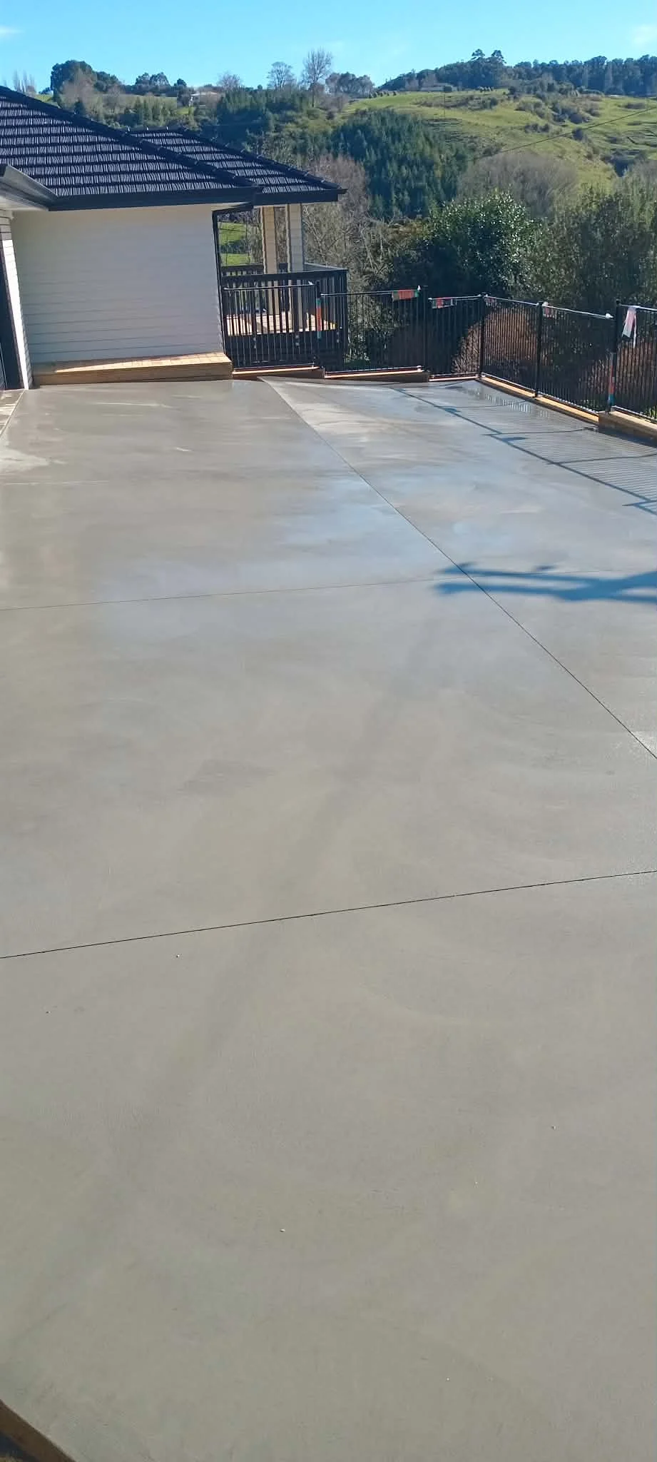 View of a spacious, newly poured concrete patio with a black metal fence and a house with white siding and a black tiled roof. In the background, rolling green hills and trees under a clear blue sky.