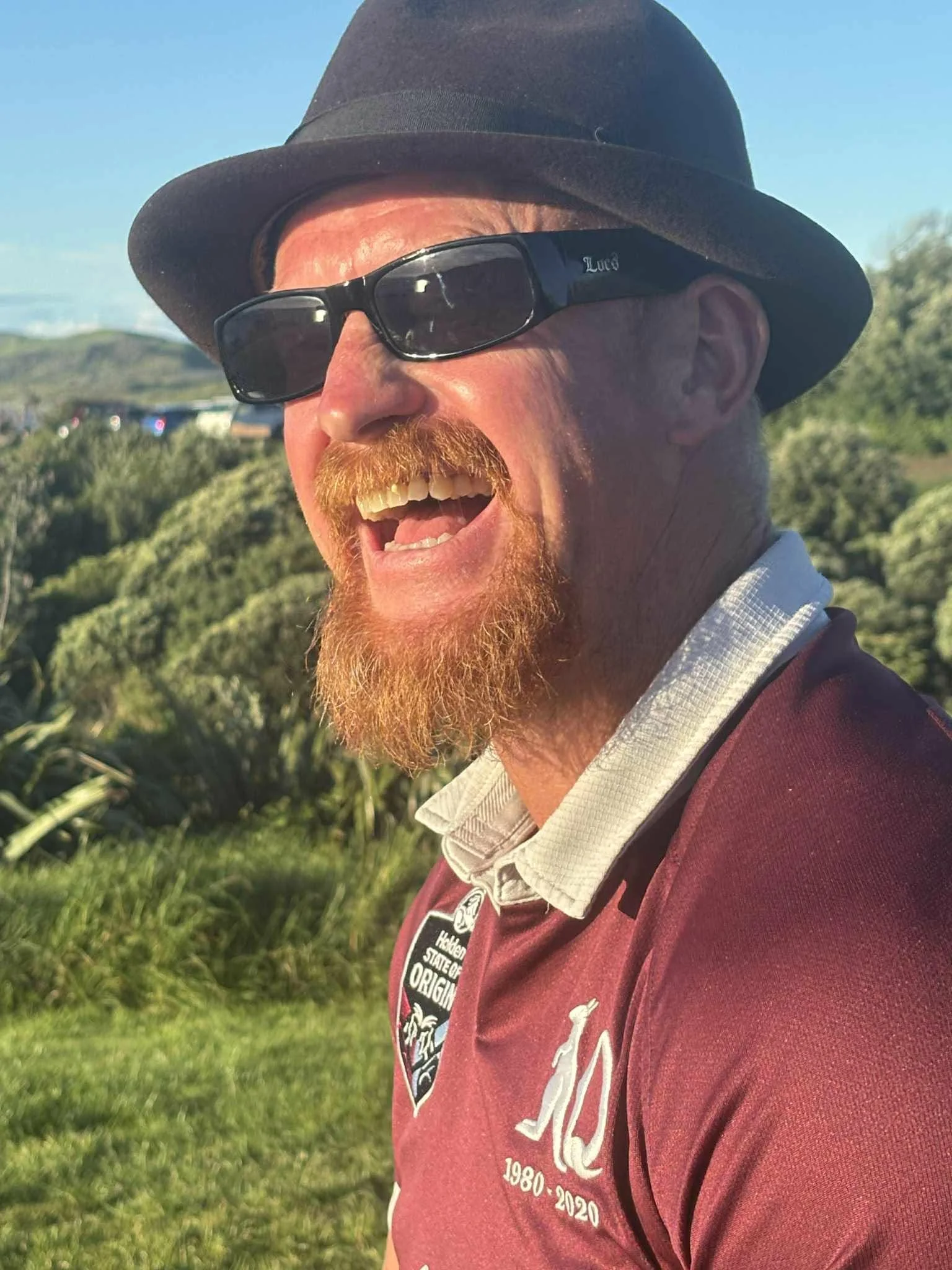 A man with a red beard and mustache wearing a black fedora, sunglasses, a collared shirt, and a maroon sports jersey with patches, smiling outdoors in a green, sunny environment.