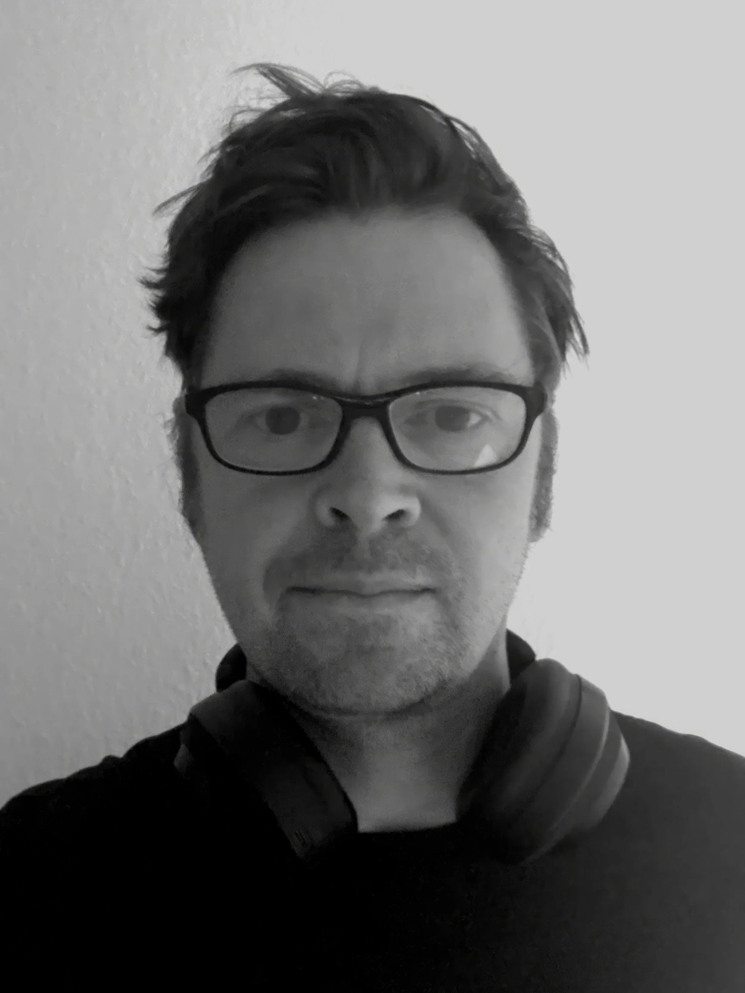 Black and white photo of a man with glasses, slightly tousled hair, and a neutral expression. He is wearing a dark shirt and has headphones around his neck, standing against a plain wall.