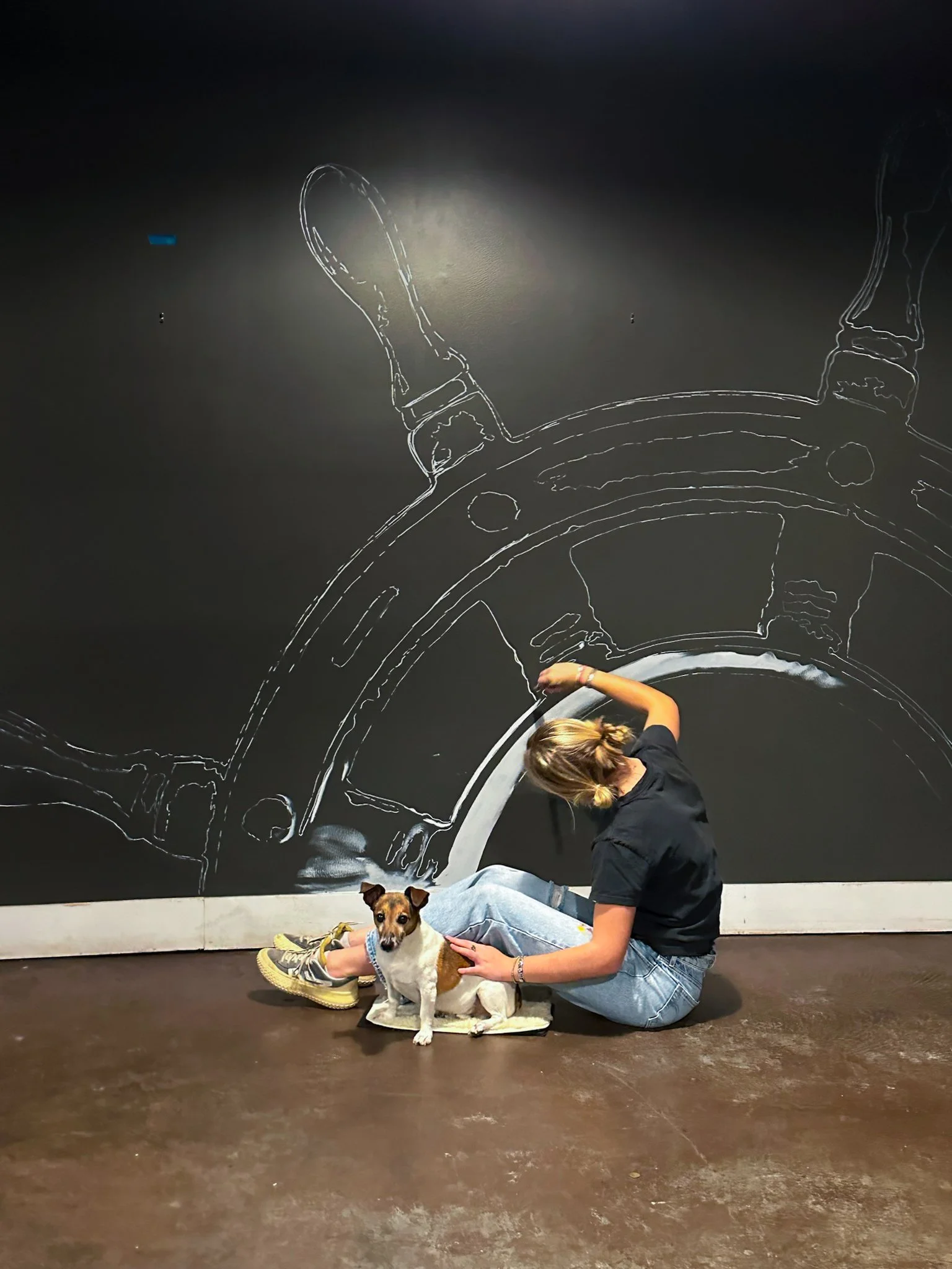 A woman with a dog sitting on a mat in front of a black wall with a white chalk drawing of a ship's steering wheel.