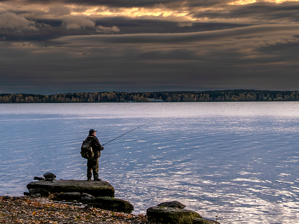 A person fishing on a rock by the water during sunset with a cloudy sky and forest in the background.