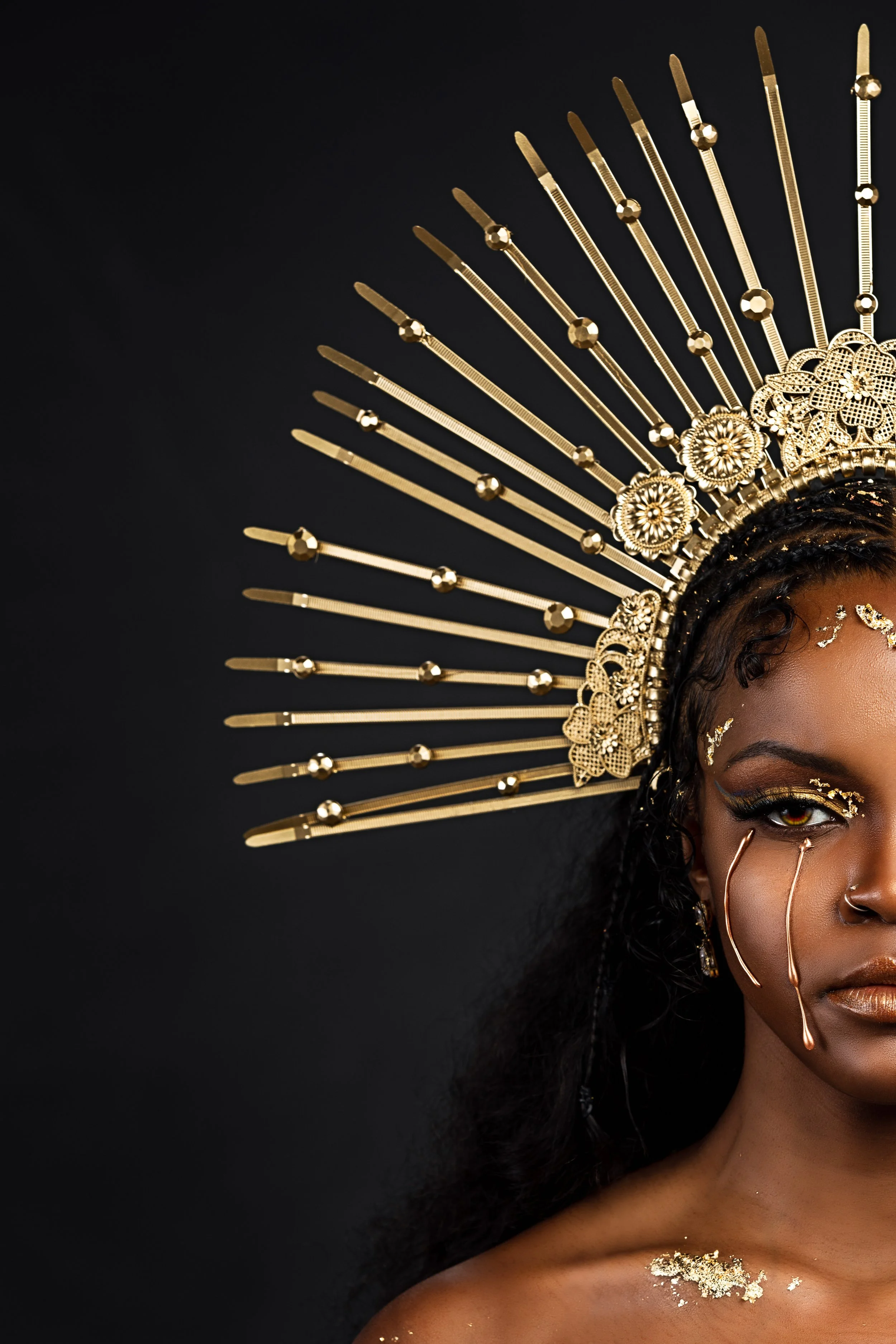 A woman with gold makeup and gold leaf accents on her face and shoulder, wearing a large ornate gold headpiece with rays extending outward against a black background.