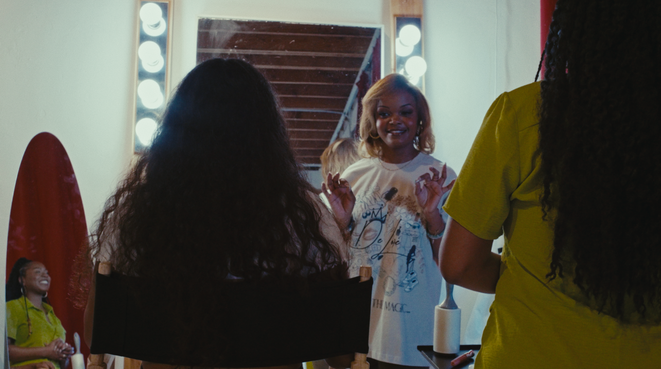 Three women in a backstage area, one with blonde hair and a white graphic t-shirt, another with black curly hair in a yellow t-shirt, and a third with long curly hair, are preparing for a show or photoshoot. A mirror with lights and a red surfboard are visible in the background.
