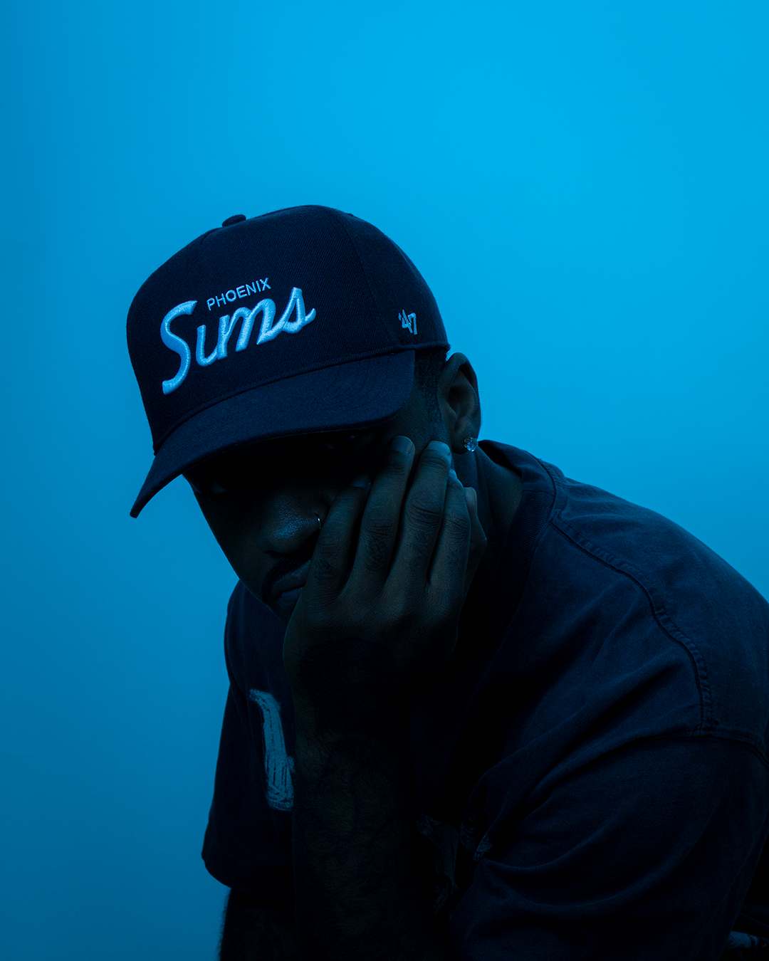 A person wearing a black cap with the words "PHOENIX Suma" and a small logo, resting their face in their hand against a blue background.
