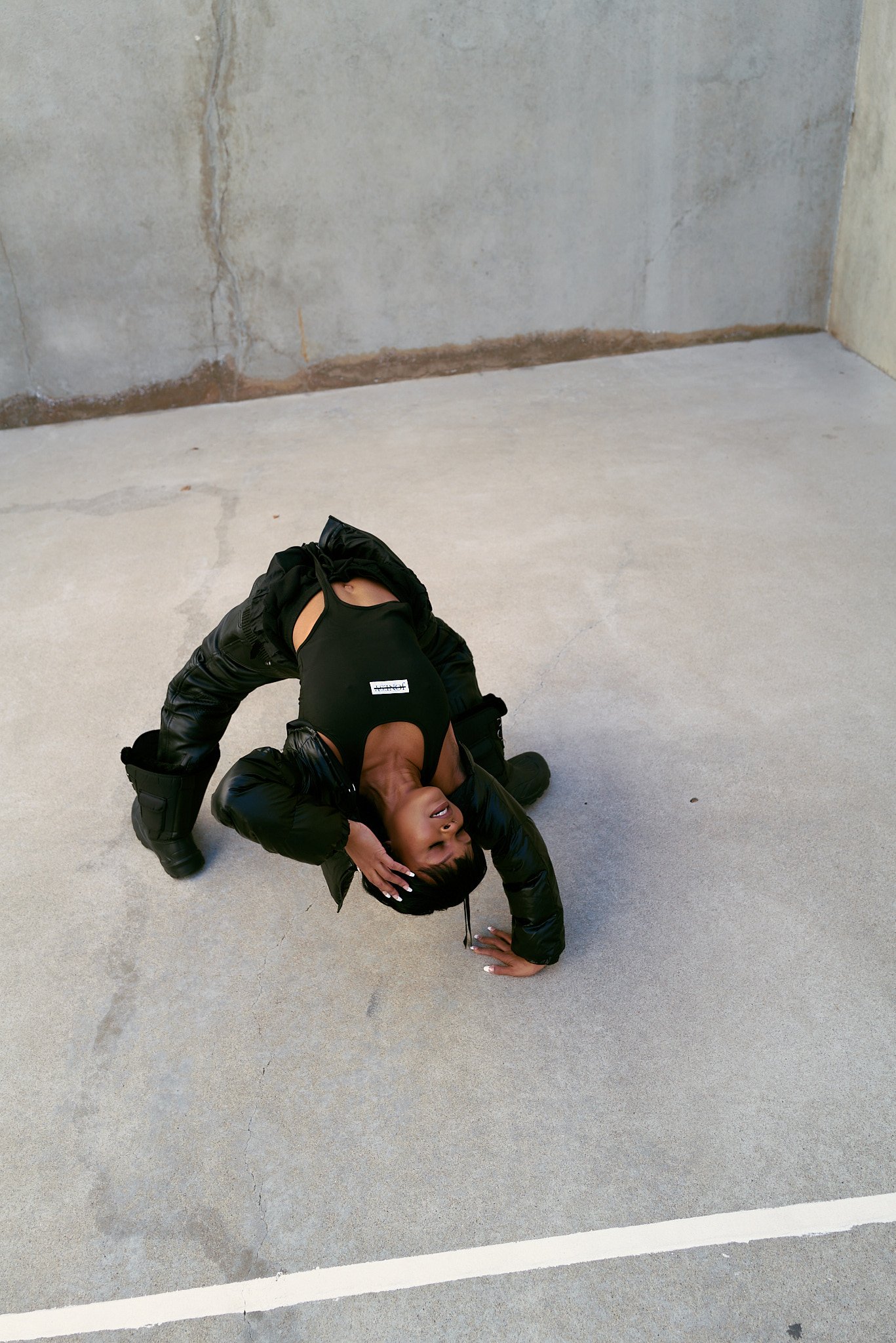 A woman in black clothing and black boots is posed in a contorted position on a concrete floor, with her head resting on one hand and her mouth open.
