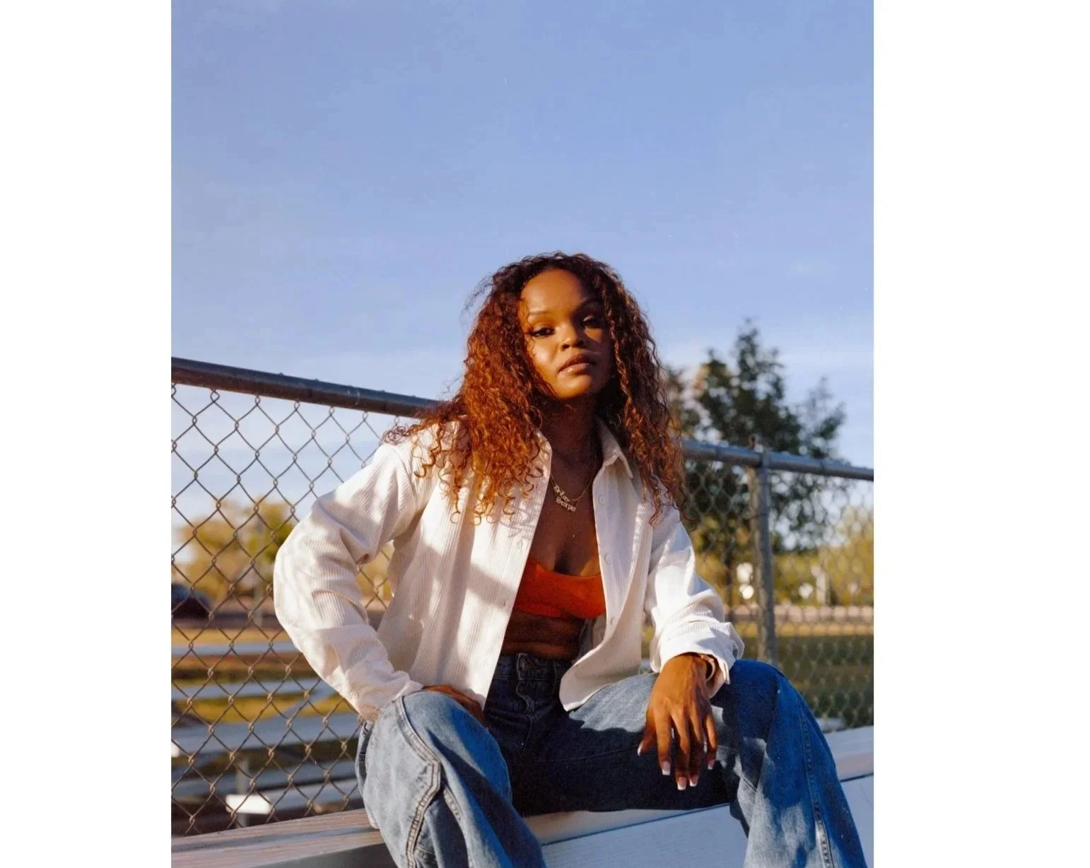 A young woman with curly hair sitting on a bench in front of a chain-link fence during sunset, wearing a white jacket, orange top, and blue jeans.