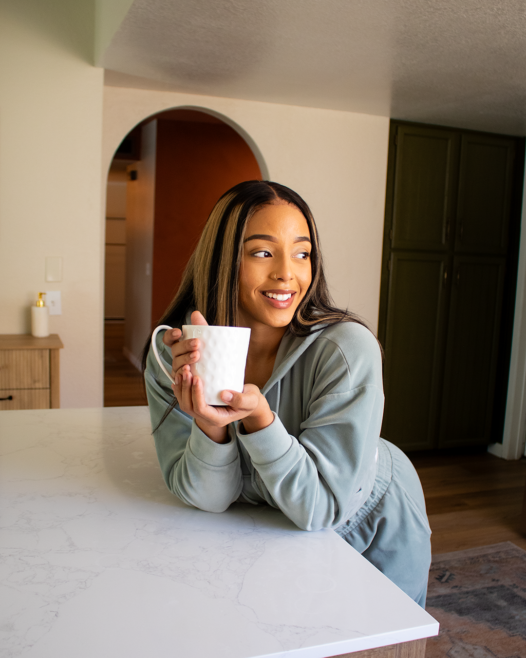 A woman with long hair holds a white mug and smiling while leaning on a kitchen counter.