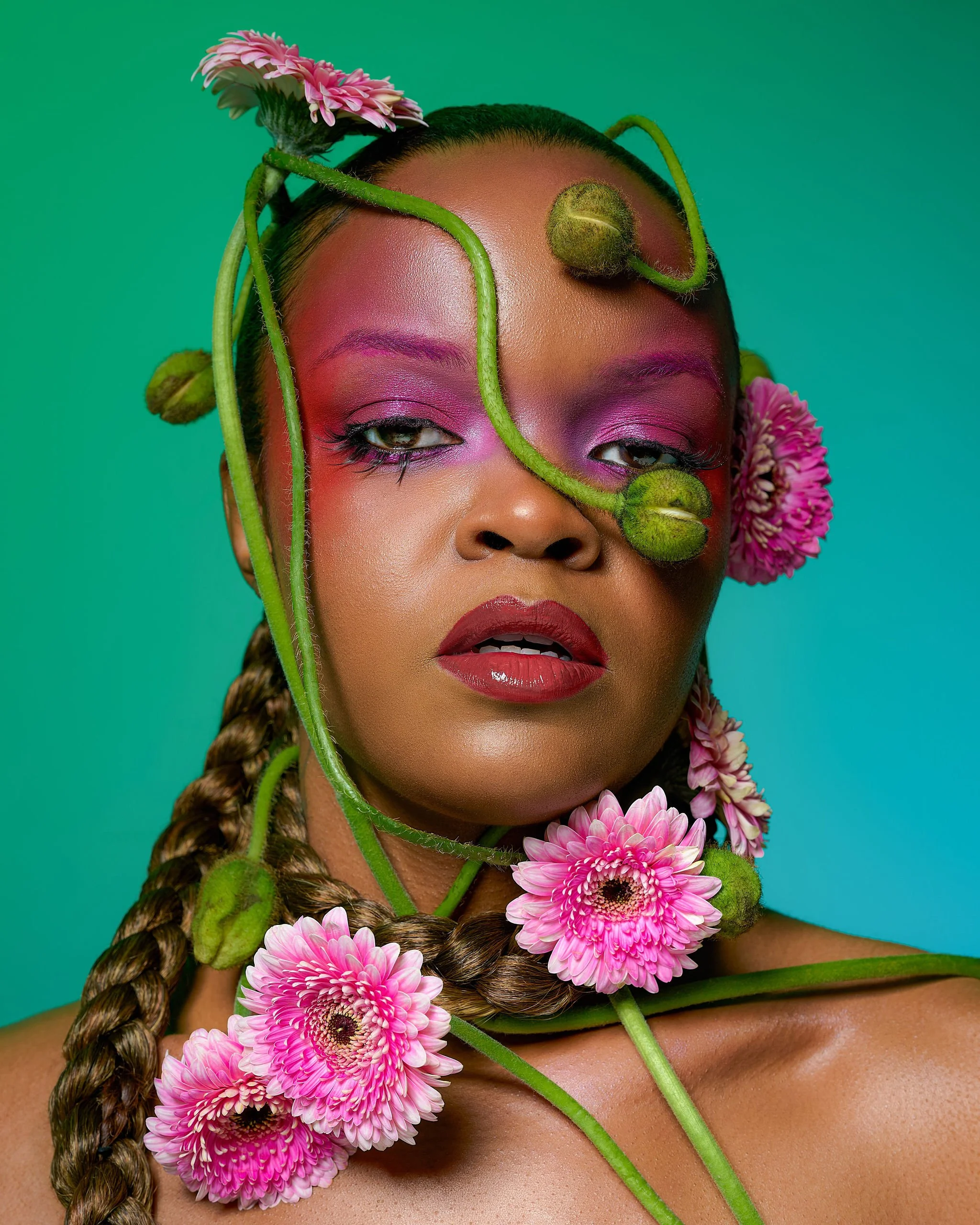Portrait of a woman with pink and purple makeup, braided hair, and pink flowers and green plant stems around her face and neck against a teal background.