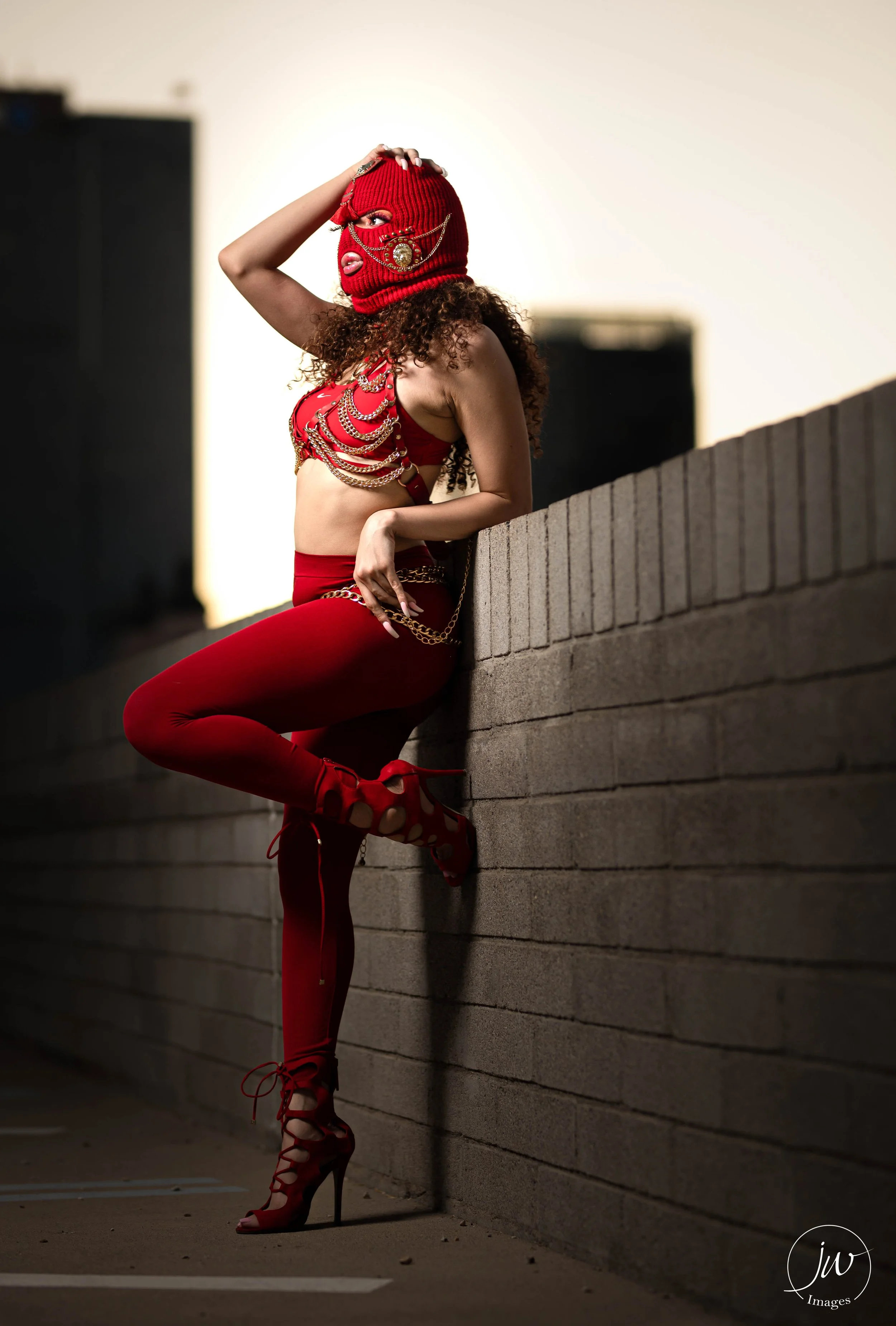 A woman dressed in red fashion with chains, wearing a red knit ski mask with embellishments, posing against a brick wall on an urban rooftop at sunset.