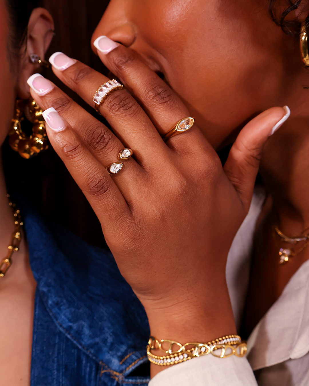 Close-up of a woman wearing multiple gold and diamond rings, earrings, a bracelet, and a necklace, with her hand near her face.