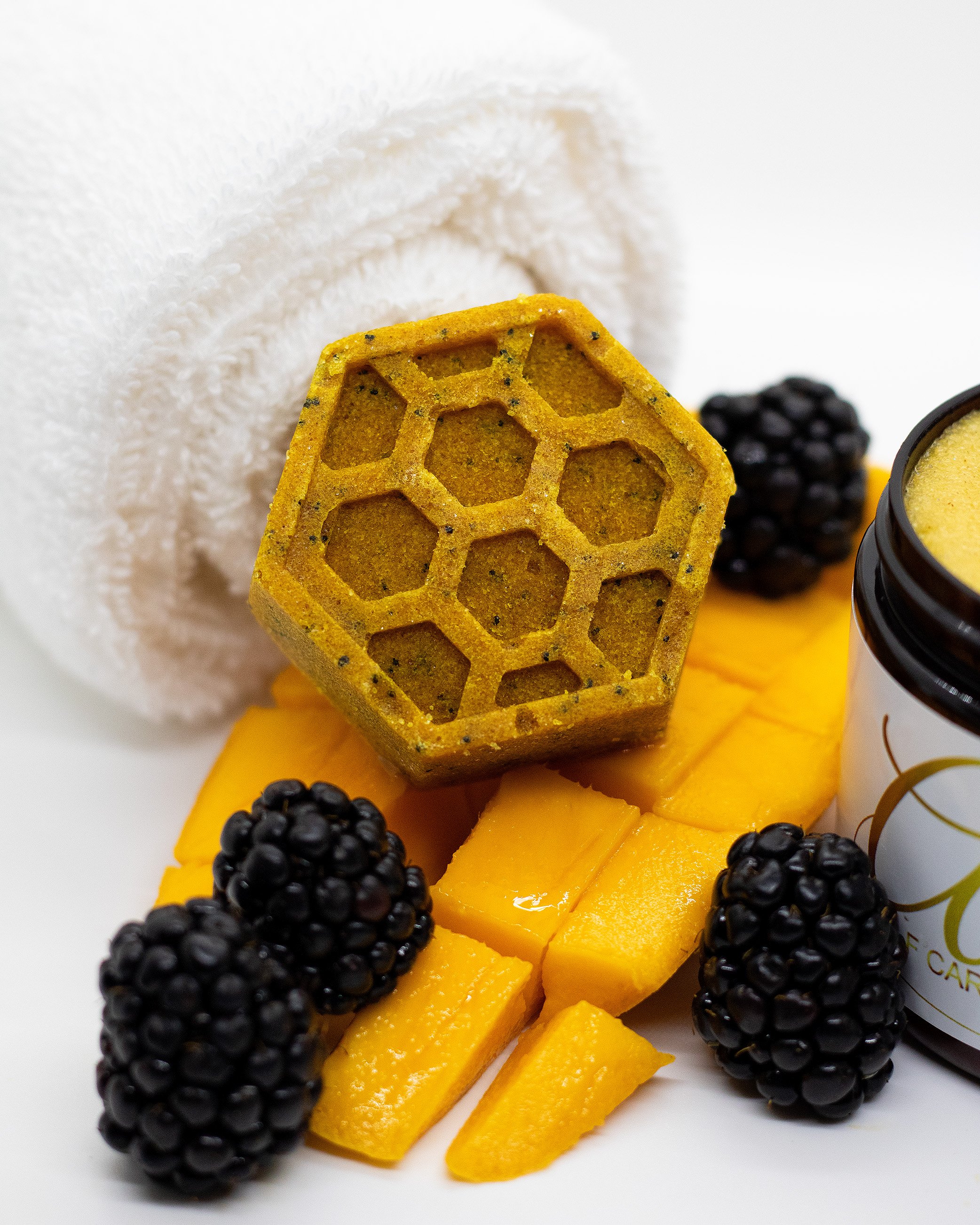 A white towel, honeycomb-shaped soap, slices of mango, blackberries, and a jar of honey on a white surface.