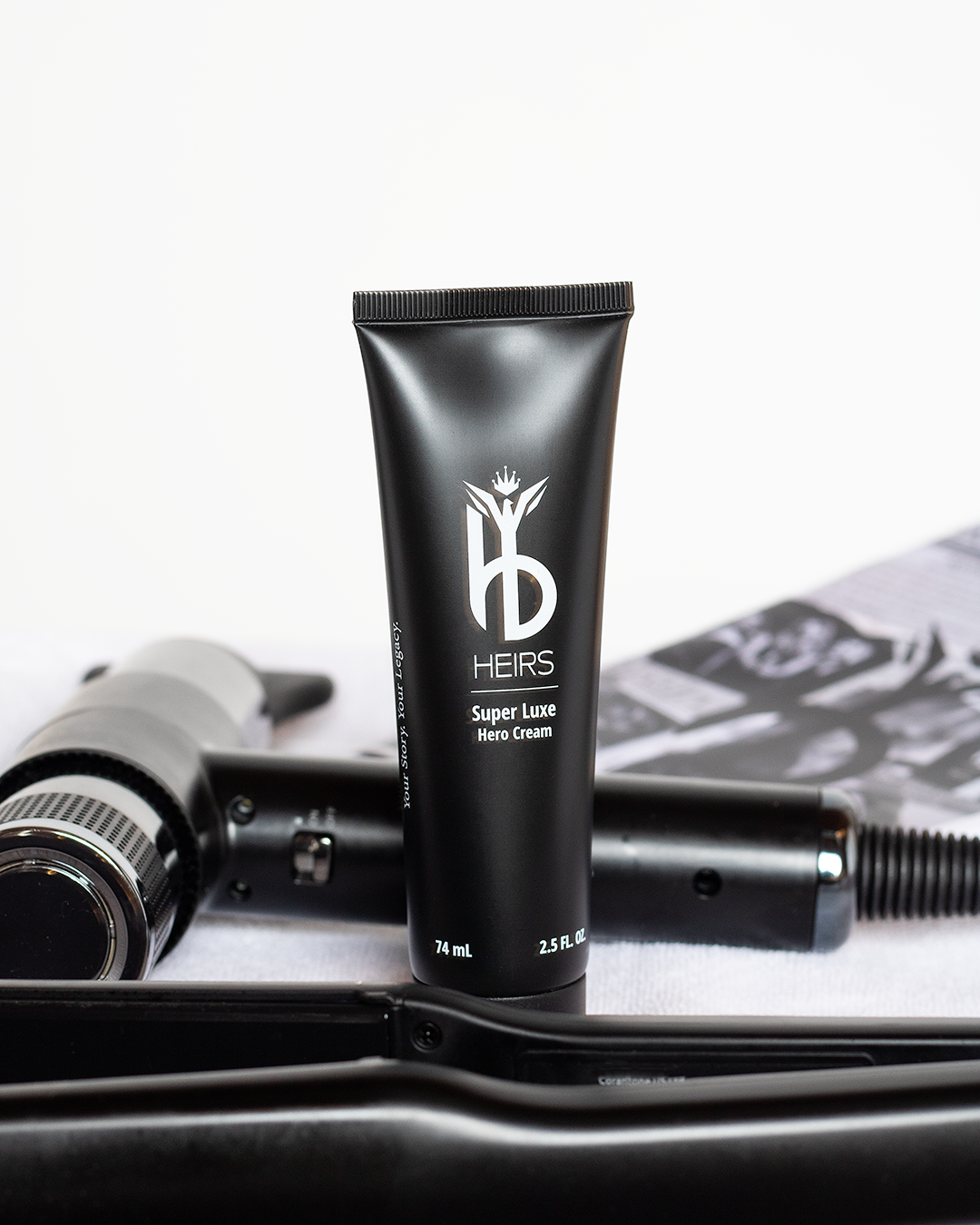 A black tube labeled 'HEIRS Super Luxe Hero Cream' stands upright on a surface, with a hairdryer and magazine in the background.