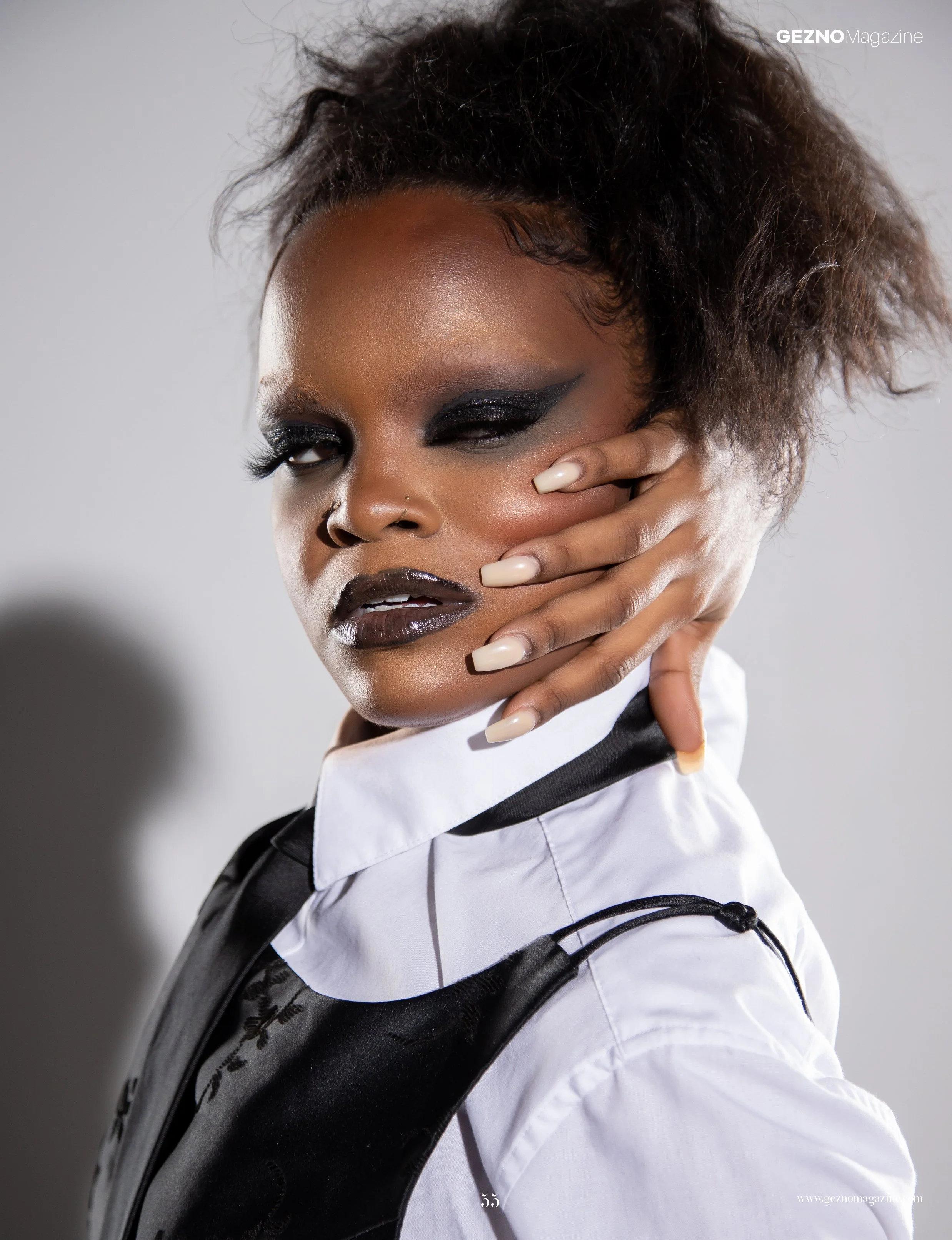 A woman with dark makeup and curly hair, wearing a black and white outfit, posing with her hand near her face against a plain background.